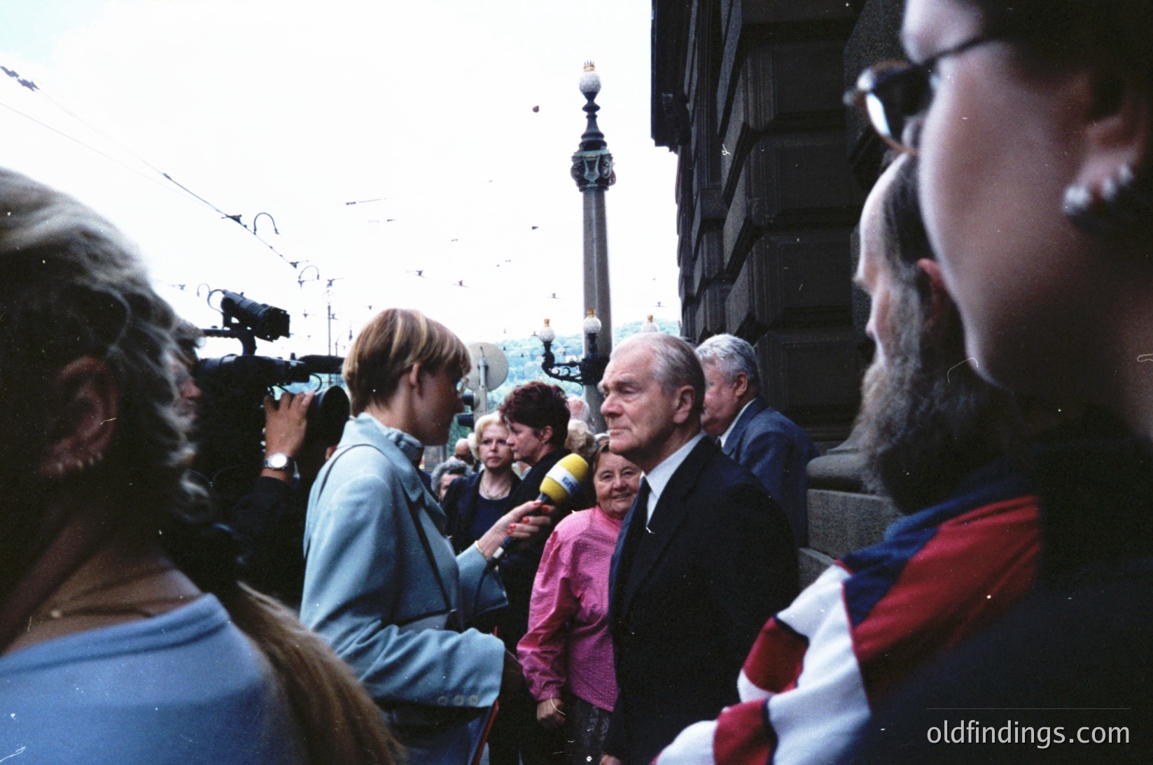 1970s-era public event near Nelson’s Column, London. Elderly man in suit speaking to crowd, surrounded by media (microphone, camera crew). Crowd wears 70s fashion (blazers, scarves). Urban setting with historic architecture and street lamps.