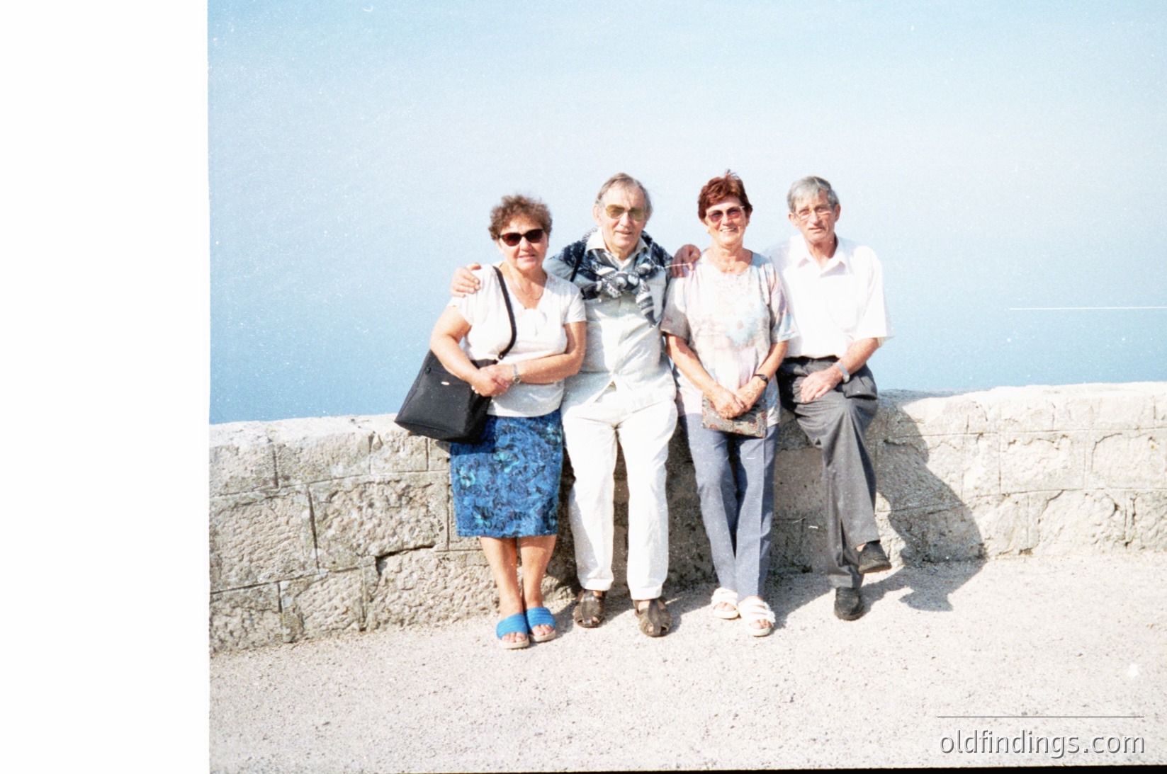 Four adults pose on a stone seawall by a calm sea, mid-1980s–1990s. Light-colored stone wall, neutral tones, and vintage camera grain suggest a seaside location. Clothing: patterned skirt, sunglasses, striped shirt, jeans, and a handbag.