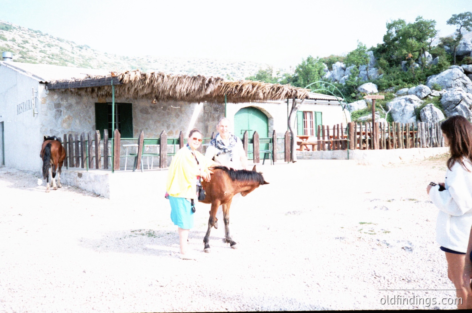 Rural courtyard with thatched-roof structures and stone walls. A woman in a yellow top and blue shorts leads a young brown horse, while another horse stands in the background. A person in white shorts and a white top captures the scene. Arched green-painted doorways and wooden fencing detail the rustic setting. Likely Mediterranean or Southern European rural area, mid-20th century.
