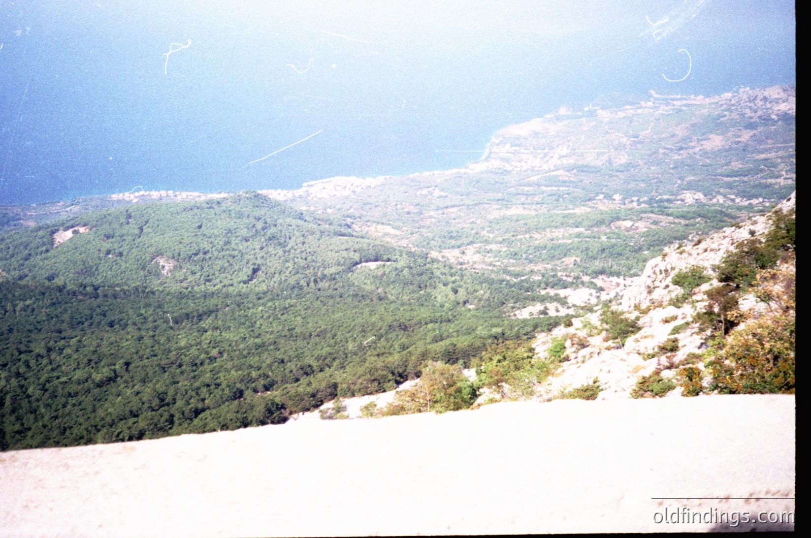Vintage aerial view of rugged alpine terrain with dense coniferous forests and rocky outcrops. Snow patches remain on higher elevations, suggesting early spring or late autumn. The hazy horizon suggests mid-20th century film grain.