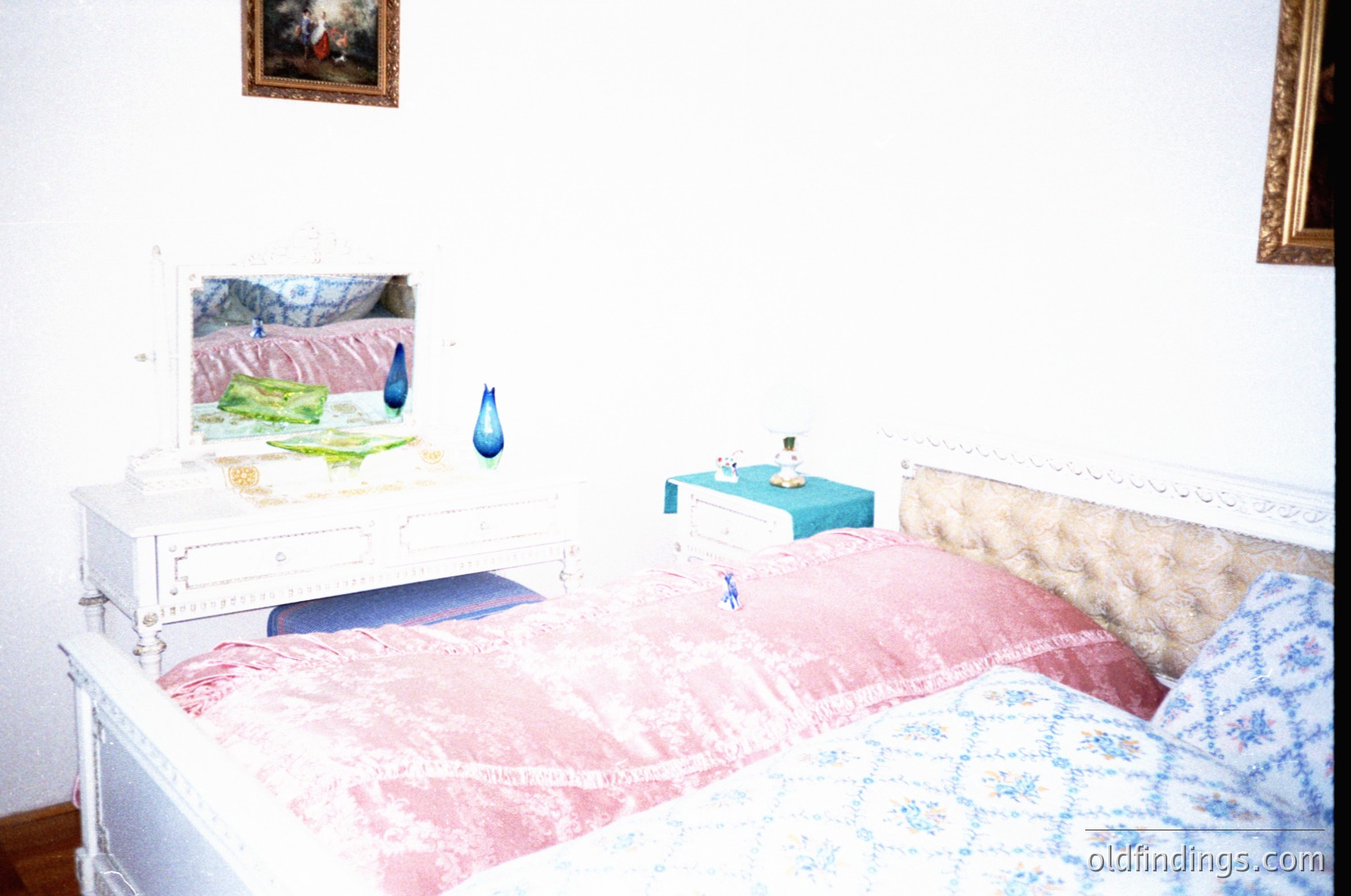 Vintage bedroom featuring a white metal-framed bed with floral-patterned bedding. A framed abstract painting (blue teardrop motif) hangs above a small nightstand with a blue ceramic vase. Wall-mounted framed artwork and a classic gilded mirror add character. Mid-century design elements suggest 1960s-70s European decor.