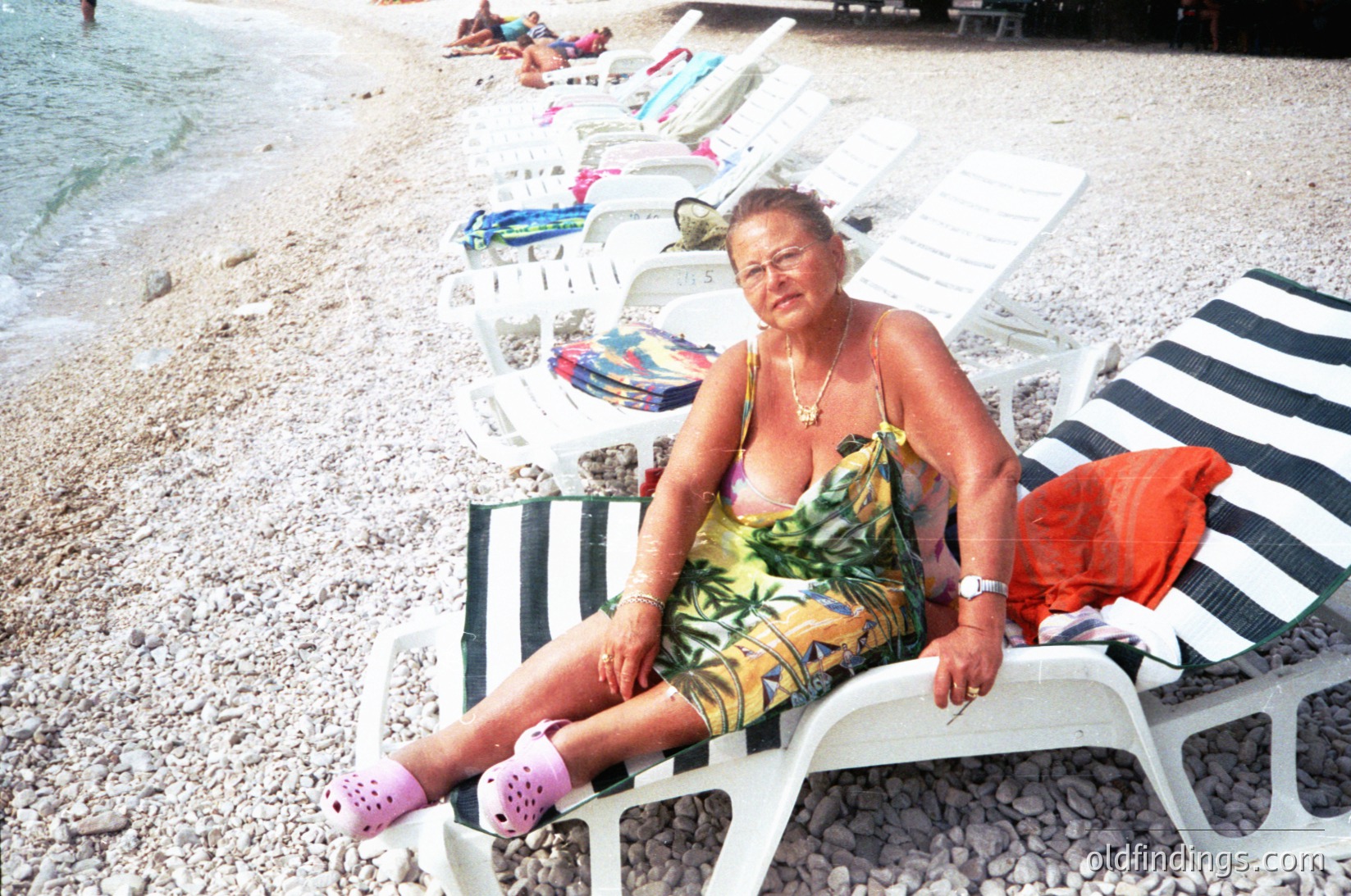 Mid-20th century seaside scene: Woman in a floral swimsuit and pink flip-flops lounges on a pebble beach, surrounded by vintage striped deck chairs. Sun-bleached towels and a red bag lie nearby. Likely Mediterranean or Eastern European coastal resort, 1960s–1980s.