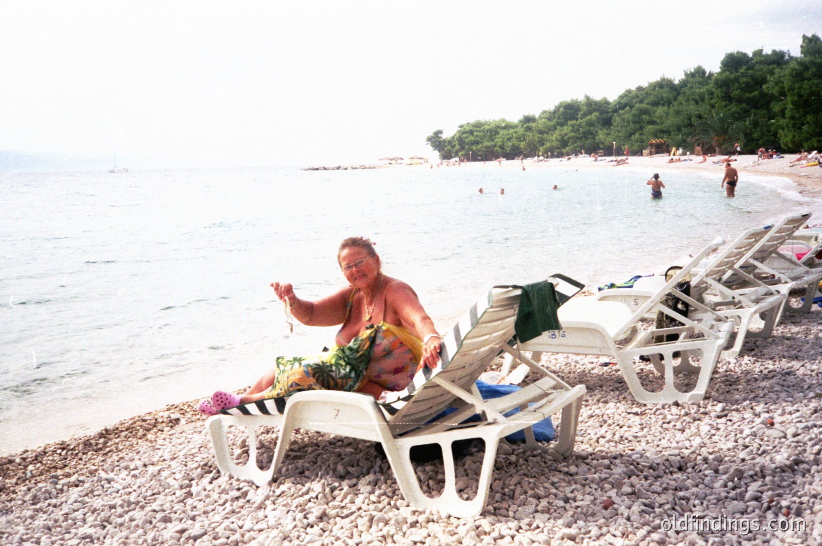A relaxed individual sits on a pebble beach in a floral sarong, holding a woven bracelet, with lounge chairs and towels in a seaside resort setting.
