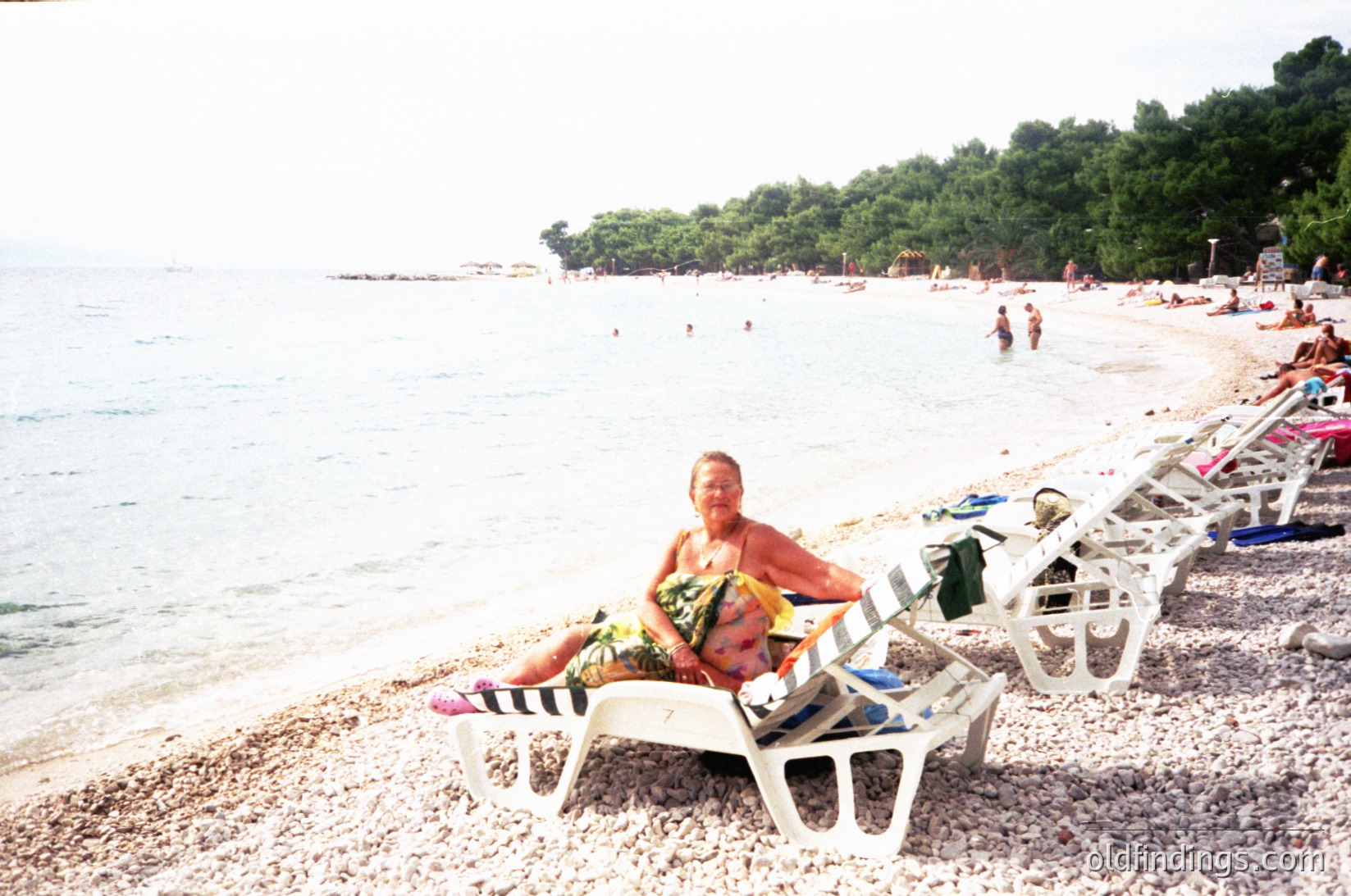 Two adults relax on vintage-style beach lounge chairs on a pebble shore, holding a striped towel. The turquoise sea and forested coastline in background suggest a Mediterranean beach setting. Crowded with sunbathers and swimmers, likely a popular seaside destination. Style evokes 1970s-1980s beach culture.