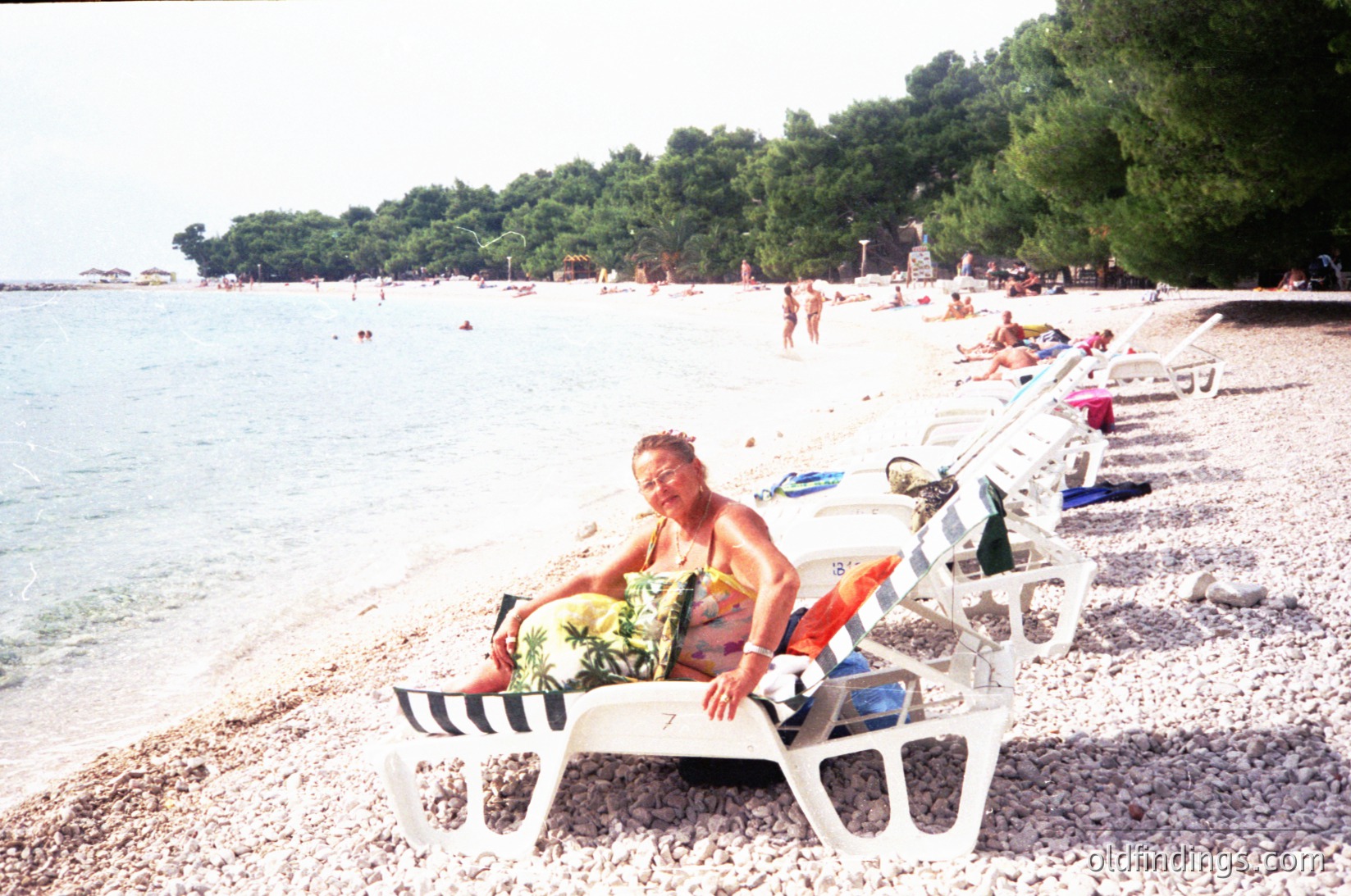 Vintage seaside scene with a relaxed individual reclining on a classic striped beach chair, holding a fruit platter. Pebble beach lined with additional chairs and sunbathers. Lush green hills and calm waters in background. Likely Mediterranean coastal area, mid-20th century.