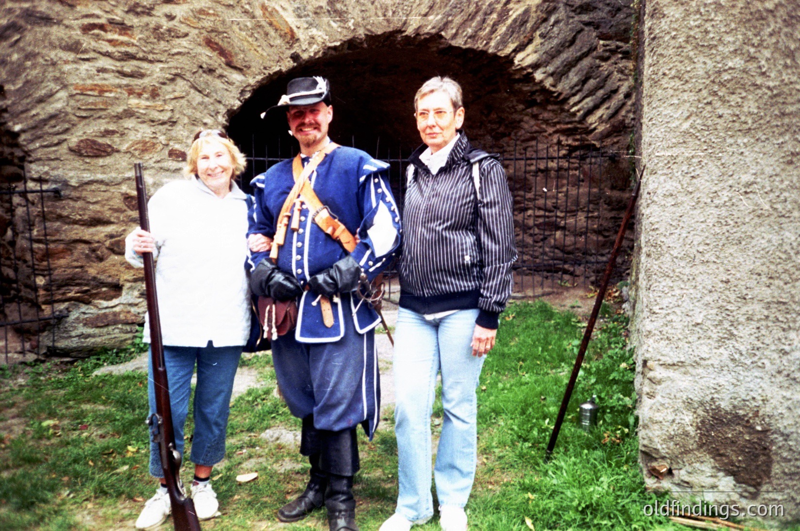 Historic reenactment scene featuring three individuals: two women in casual 1990s attire and a man in mid-19th century Prussian/German military uniform. Stone archway and fortress wall suggest a European military site. Ideal for historical education or stock imagery.