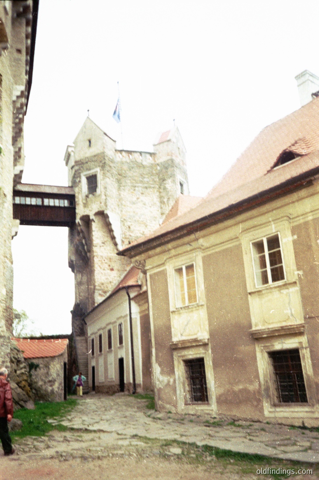 Medieval courtyard flanked by weathered stone buildings with arched windows and tiled roofs. Central tower features a flagpole and crenellated top. Cobblestone path leads to a wooden bridge over a narrow moat. Likely Eastern European castle complex, possibly or .
