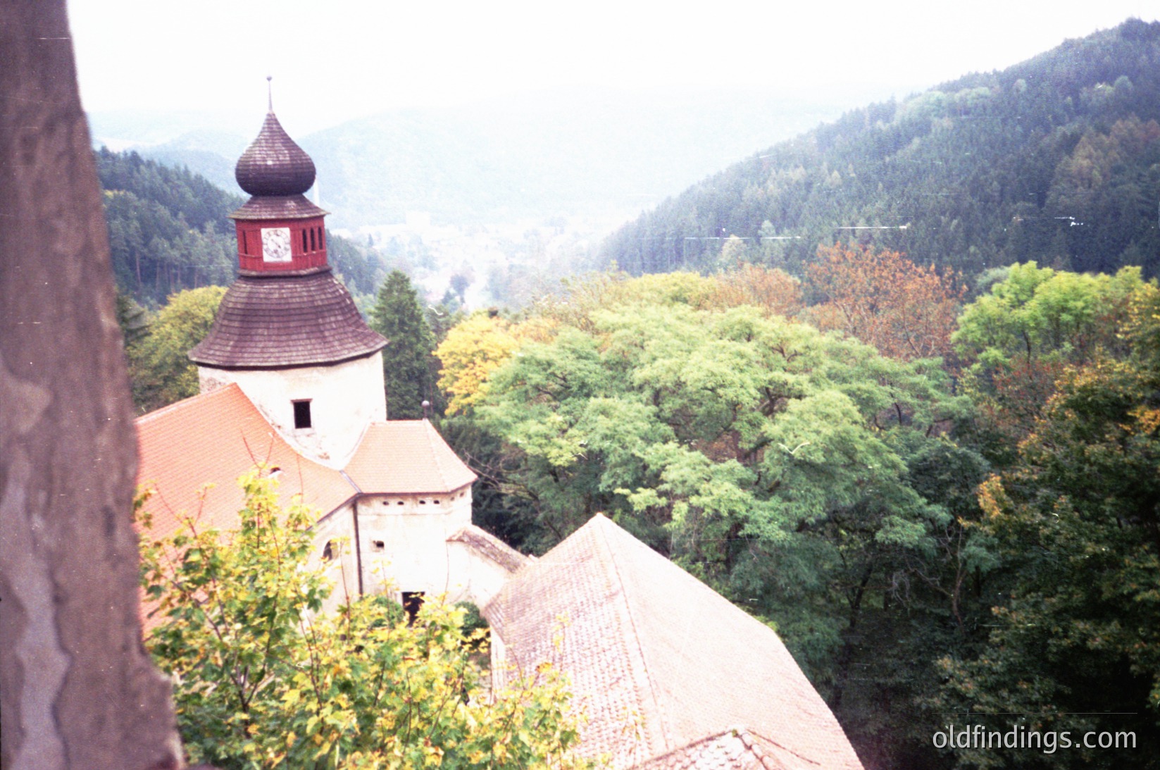 Historic castle tower with red-tiled conical roof and clock, surrounded by dense autumn foliage. Misty mountain backdrop suggests Eastern European architecture, likely 19th–early 20th century. Ideal for heritage, travel, and scenic stock imagery.