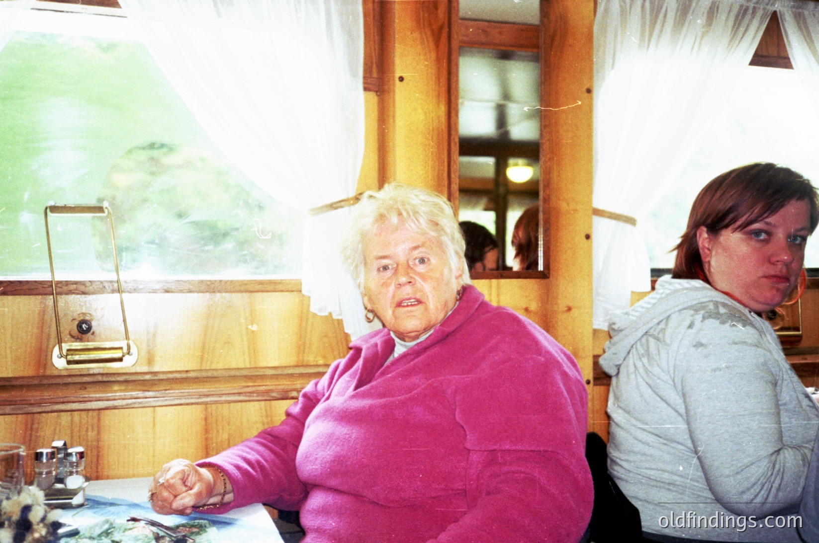 Elderly woman in a pink sweater sits at a wooden table in a rustic indoor setting, holding a small object. A younger woman in a gray hoodie stands behind her. Reflections in a mirror reveal a blurred outdoor scene with trees. Likely a rural or cabin interior, late 20th century.