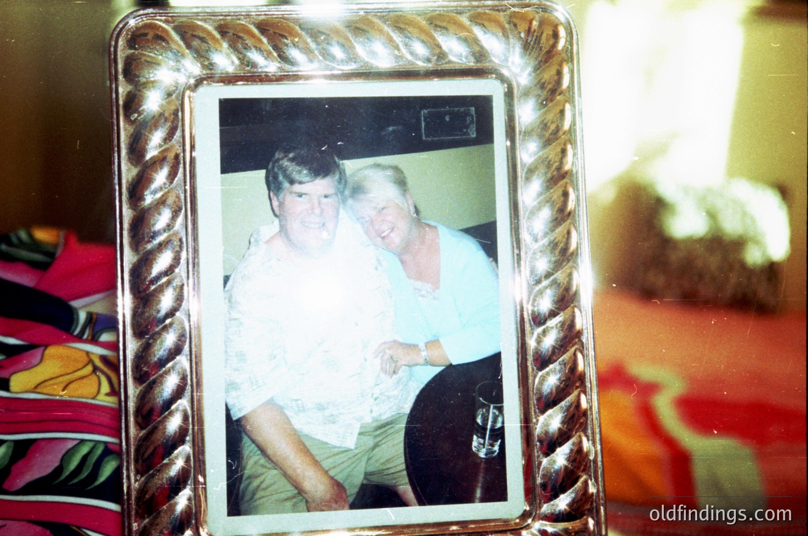 Vintage framed photo of two individuals in casual 1970s attire—man in a light shirt and khakis, woman in a floral blouse—posing indoors. Framed in a decorative metal border, set against a patterned fabric backdrop. Likely a personal keepsake or family archive.