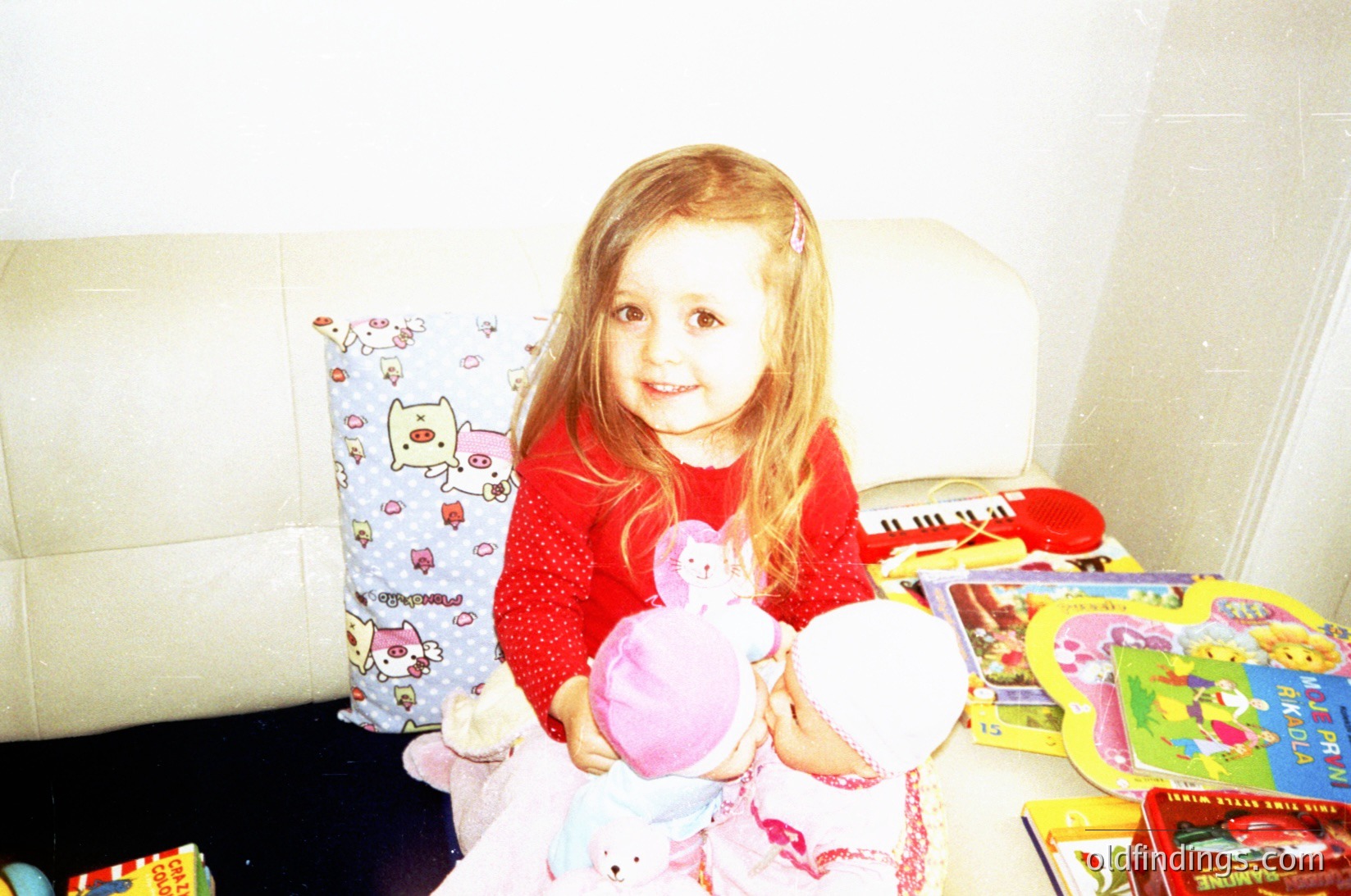 Vintage-style photo of a young girl (approx. 4-6 years) seated on a floral-patterned sofa cushion, holding a pink unicorn doll and a white teddy bear. Surrounding her are assorted toys, including a puzzle and a stuffed animal. Warm, soft lighting suggests indoor playtime, likely mid-20th century.