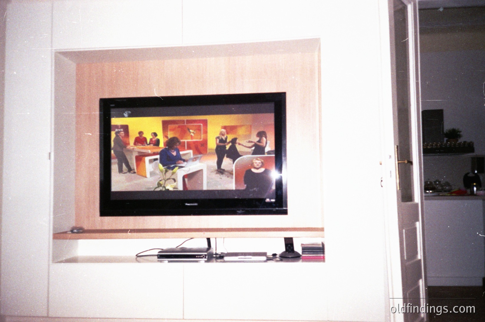 Flat-screen CRT TV mounted in recessed alcove, displaying a 1970s-style talk show with panelists and audience. Minimalist interior with built-in shelving below. Analog-era tech juxtaposed with modern minimalist decor.