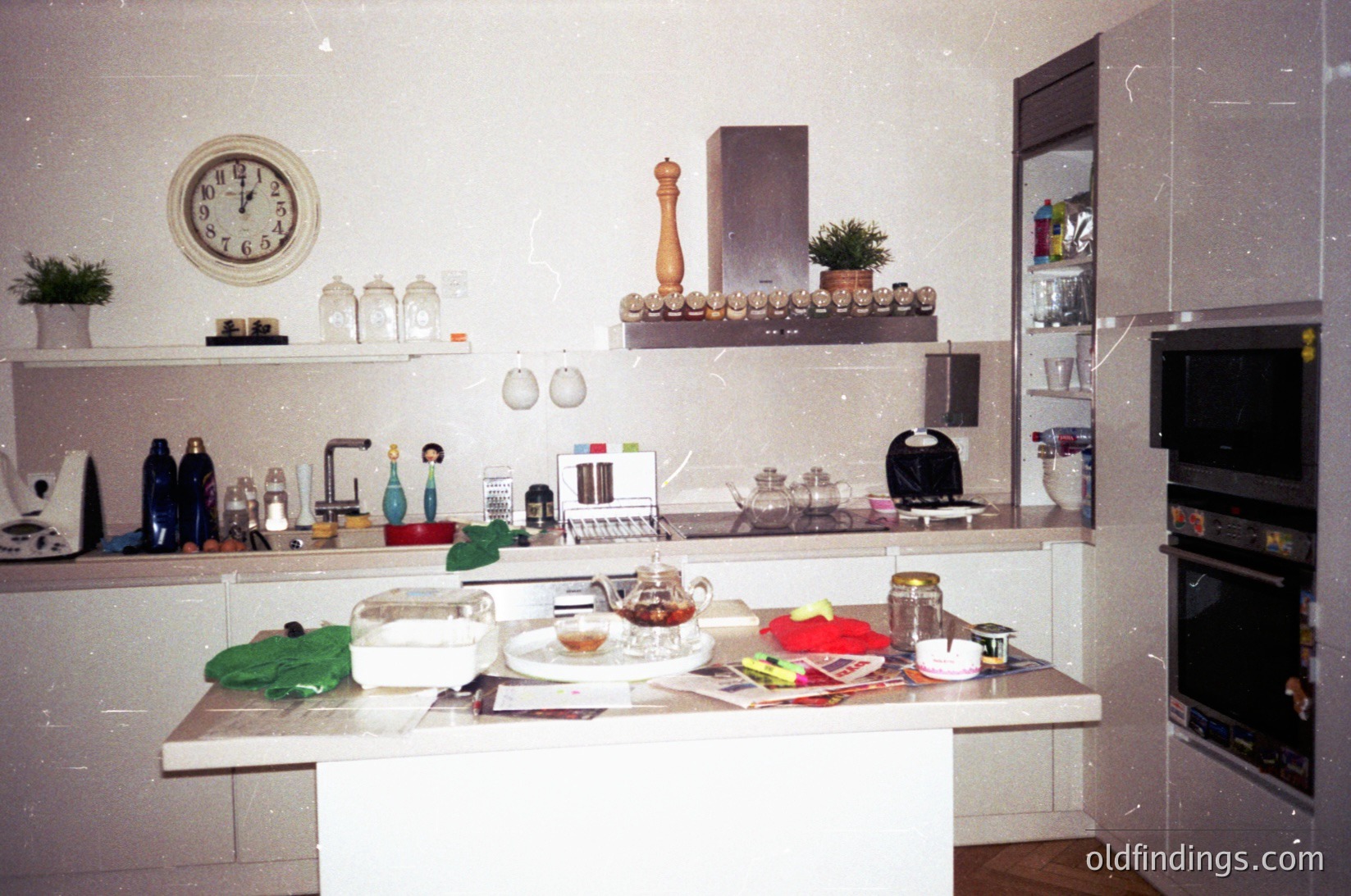 Vintage kitchen with retro white laminate countertops, featuring a built-in microwave, electric kettle, and retro clock. Decor includes ceramic figurines, glass jars, and a wooden pepper grinder. Mid-century design with built-in shelving and a built-in oven. Likely 1970s–1980s European home.
