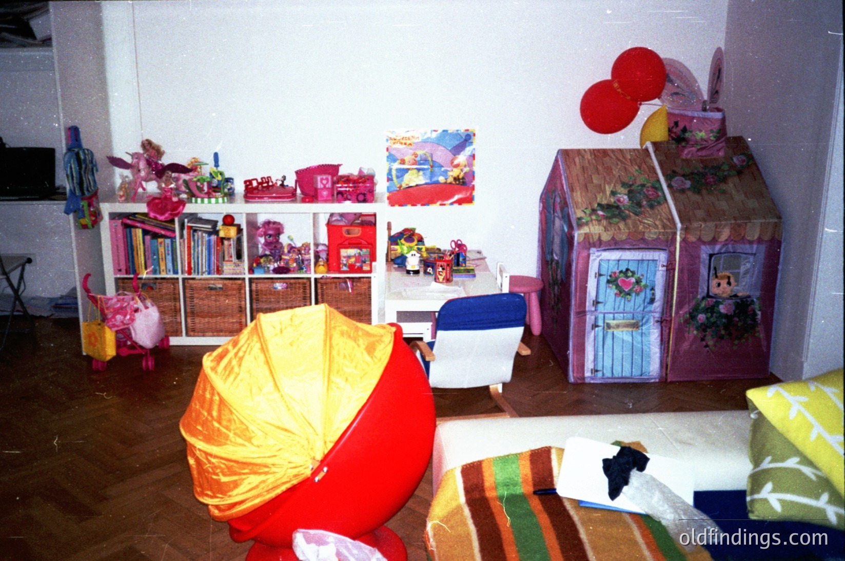 Vintage children’s playroom with mid-century play structures: cardboard house with floral details, toy shelves stocked with books, toys, and decorative bins. Bright yellow umbrella chair and striped floor cushions. Wall-mounted artwork featuring cartoon characters. Likely 1970s–1980s domestic setting.