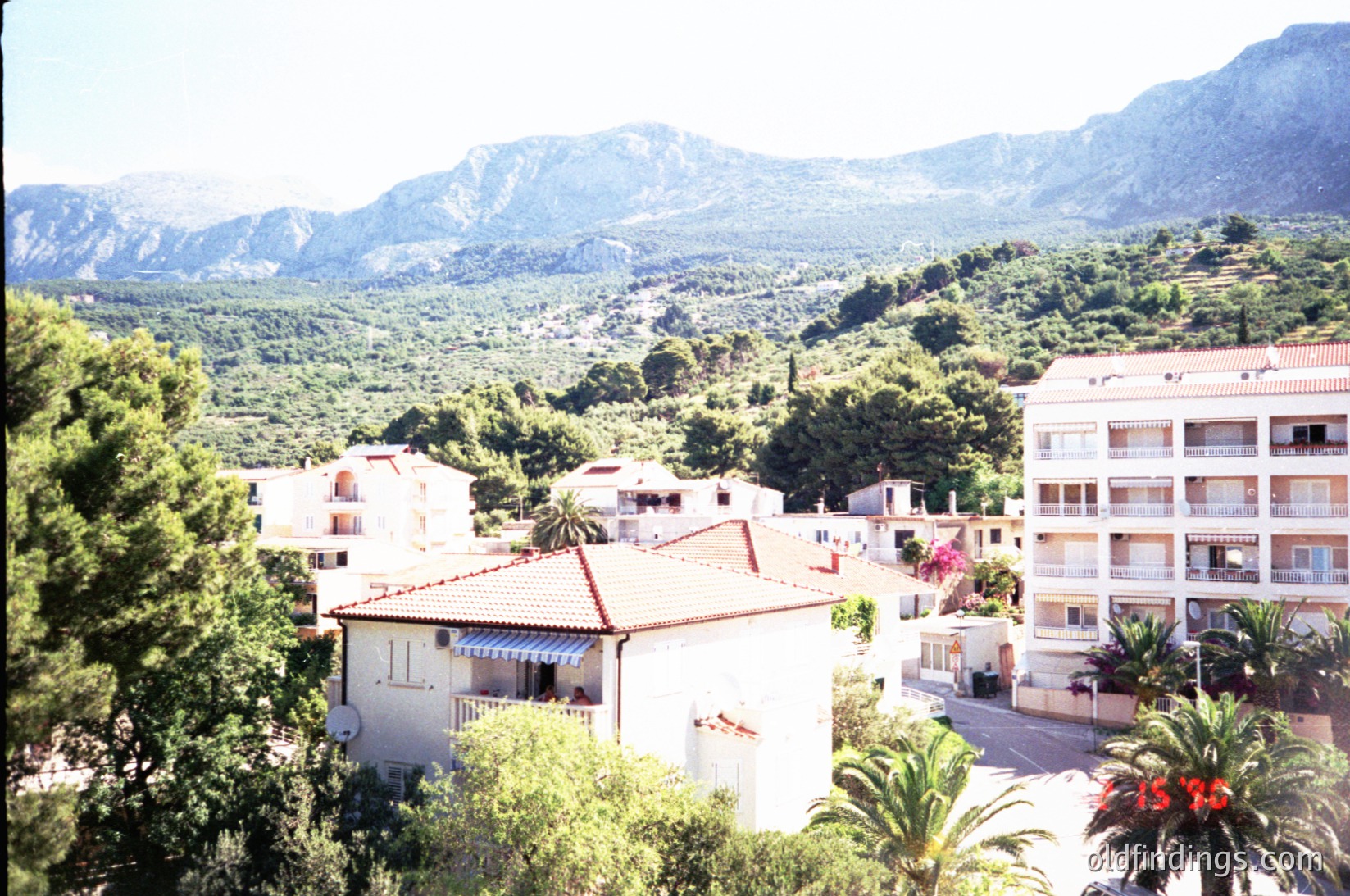 Mid-20th century Mediterranean coastal village nestled between mountains and sea. Whitewashed buildings with terracotta roofs and balconies overlook terraced hillsides. Lush greenery and palm trees frame the scene, suggesting a warm, sunny climate. Likely or , -era architecture.