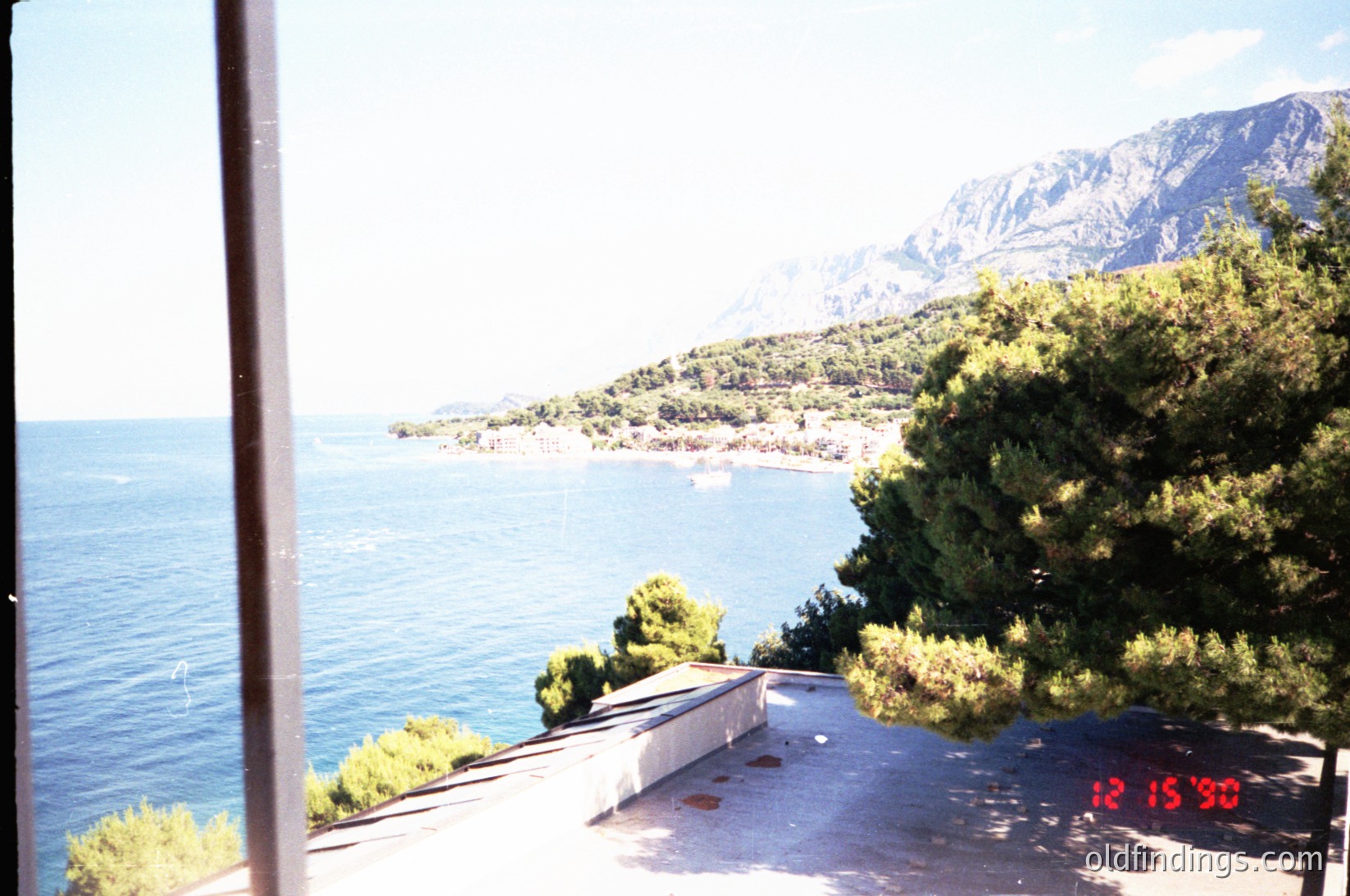 Vintage coastal view from an elevated terrace, framed by a wooden railing. Lush greenery and a concrete ledge with a small bench overlook deep blue waters meeting rugged mountains. Date stamp "12/15/90" suggests late 1990s Mediterranean or Adriatic region.