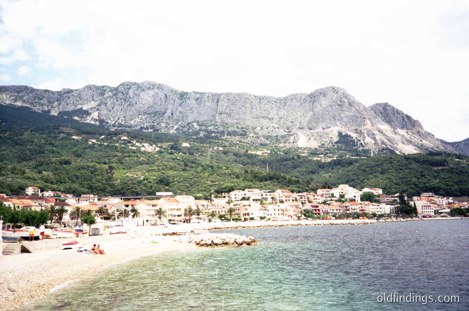 Coastal town nestled between rugged mountains and turquoise waters, featuring pebble beach with sparse sunbathers. Low-rise Mediterranean-style buildings line the shore, with palm trees and greenery dotting the landscape. Dramatic rocky cliffs frame the scene, suggesting a secluded yet scenic seaside location.