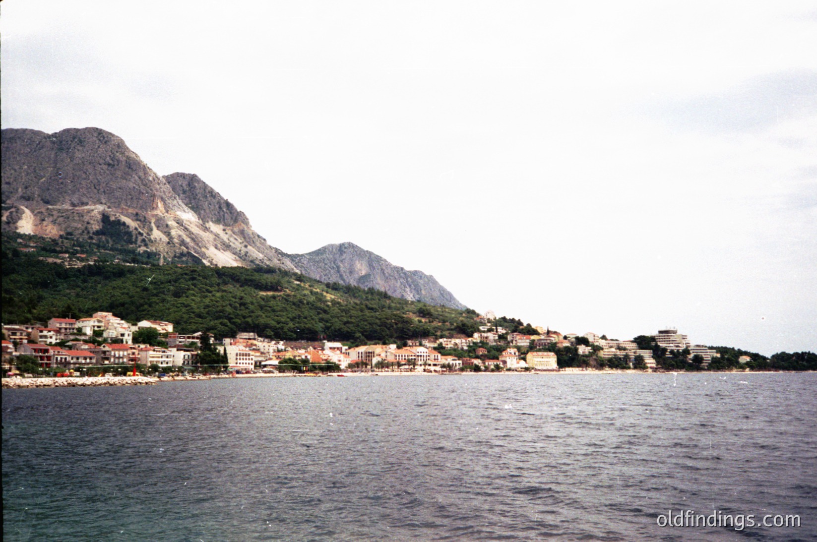 Coastal town nestled between rugged mountains and deep blue sea, likely Mediterranean. Mid-20th century architecture with low-rise buildings and flat roofs. Overcast sky suggests early morning or late afternoon lighting. Ideal for travel, real estate, or historical tourism references.