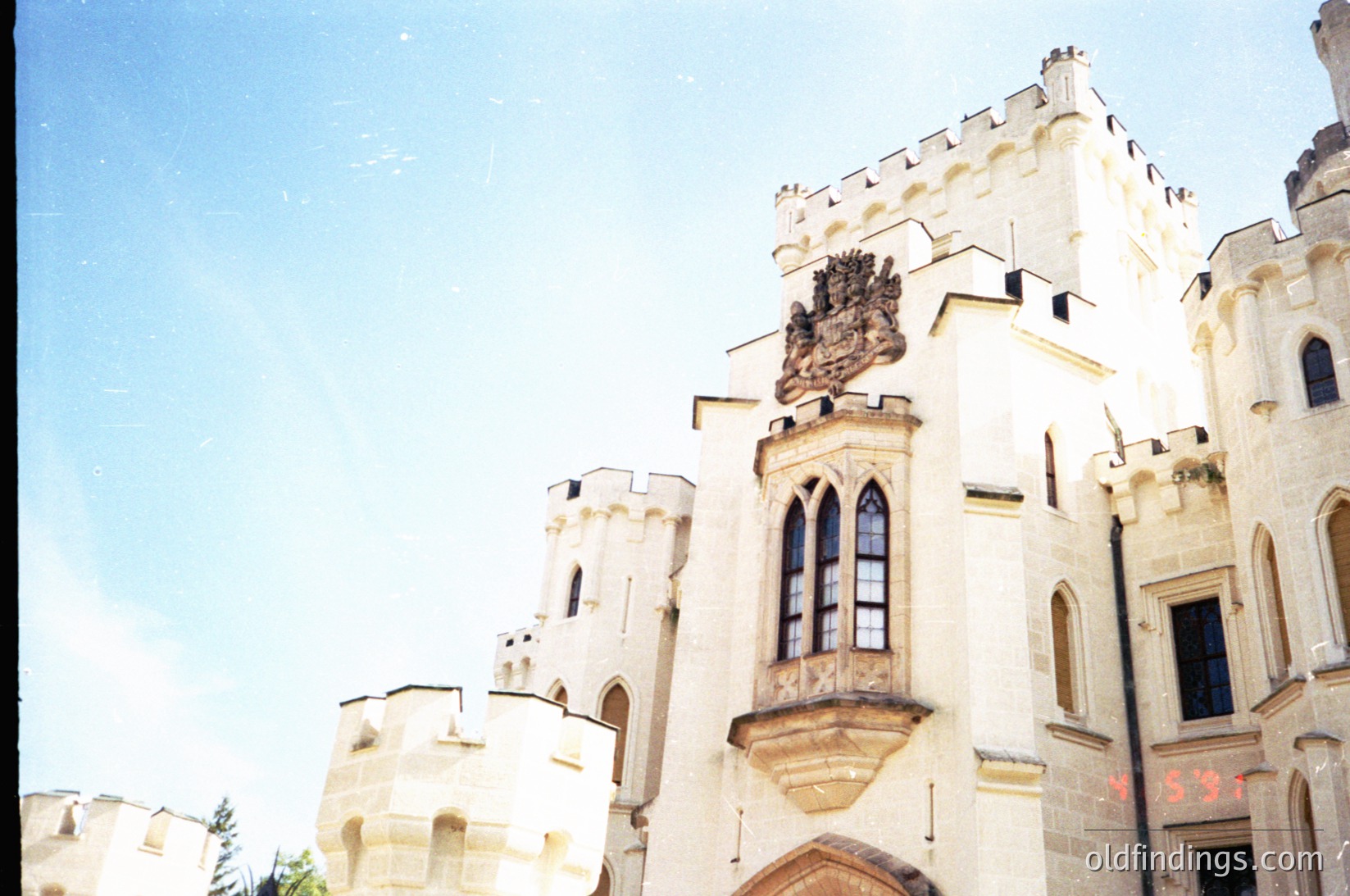 Neoclassical castle facade featuring crenellated towers, arched windows, and ornate heraldic crest. Light beige stonework with subtle weathering. Likely European, 19th–early 20th century.