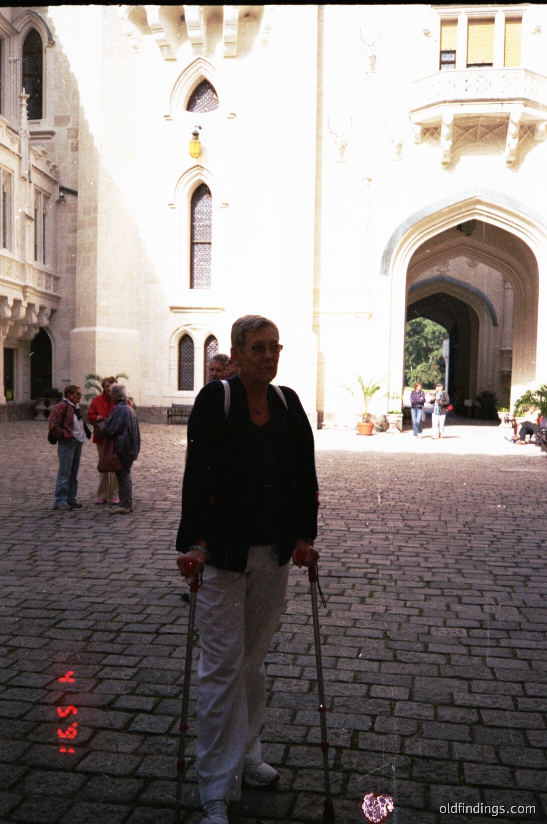 A woman in a dark sweater and light pants uses canes while walking in a historic courtyard. Gothic-style stone arches and pointed windows frame the scene, suggesting European architecture. Cobblestone pavement and a few other visitors add context. Likely late 20th century, possibly .