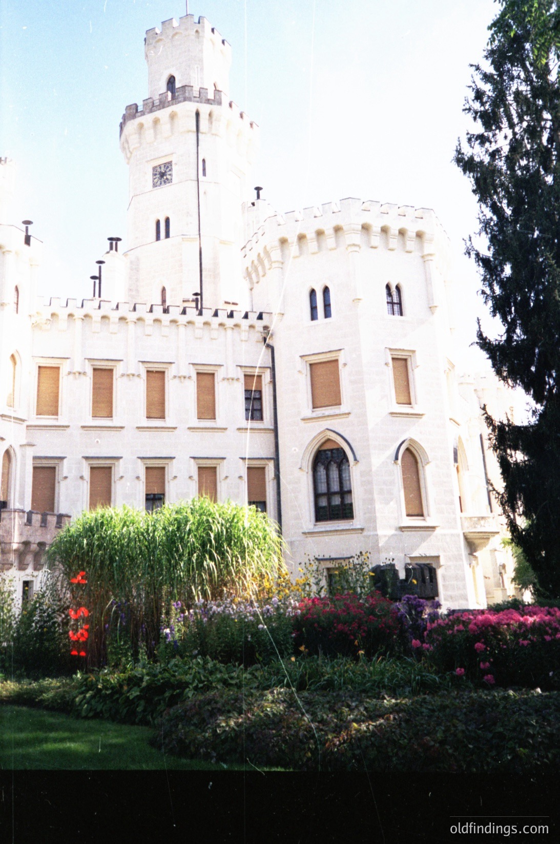 Neoclassical-style castle with crenellated towers and arched windows, set against a clear sky. Lush garden with vibrant flowers and greenery in foreground. Likely European, mid-20th century.