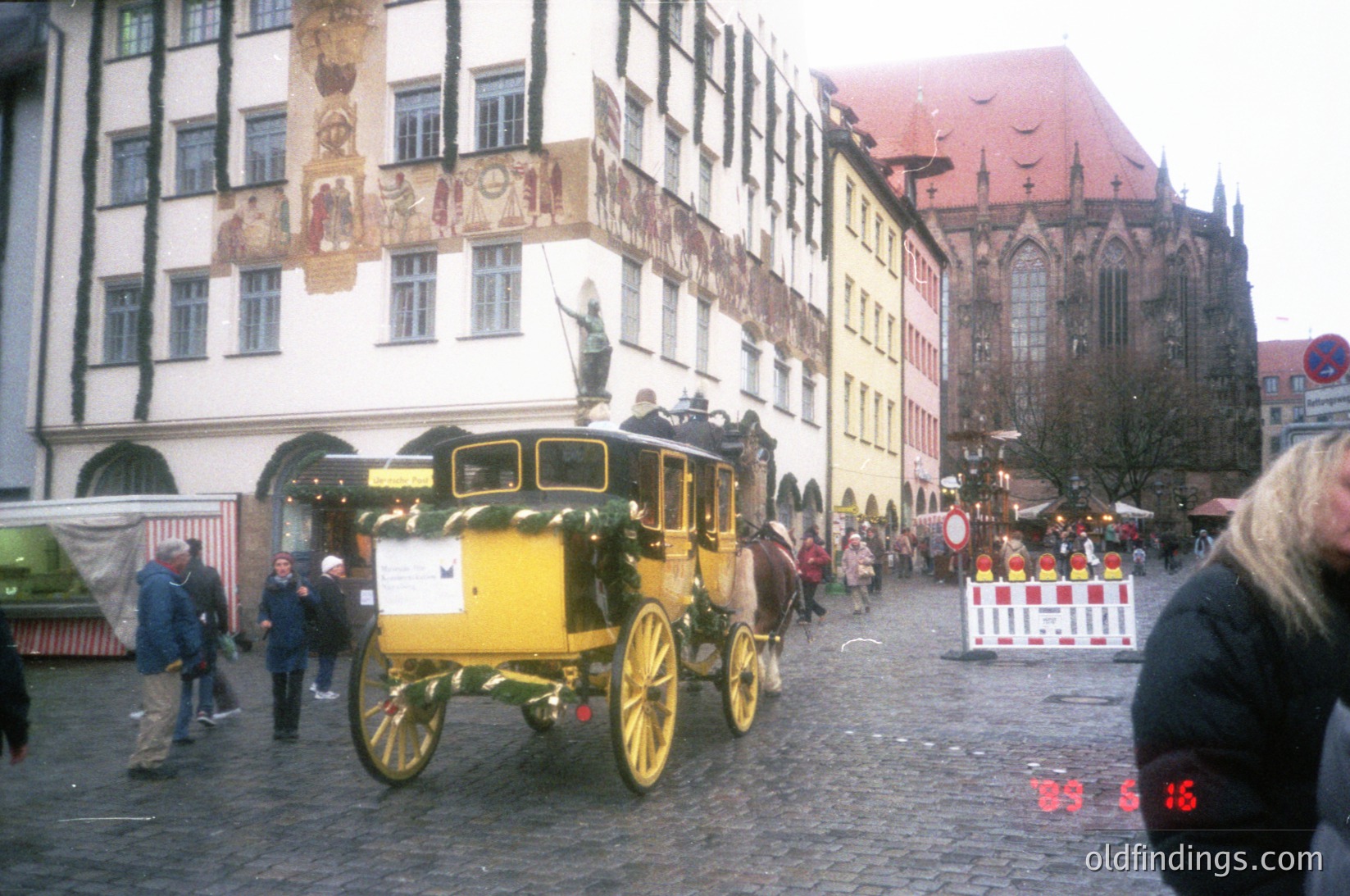Historic yellow horse-drawn carriage navigating a cobblestone square in Nuremberg, Germany. Surrounding buildings feature medieval timber-framed architecture with ornate murals. Crowds and festive stalls suggest a market or holiday event, likely late 20th century.