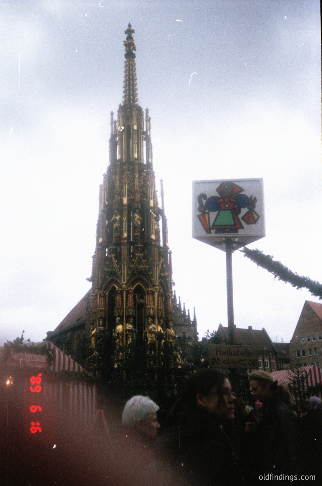 Gothic spire of the Frauenkirche in Dresden, Germany, adorned with festive garlands, likely during the 1960s or 1970s. The sign in the foreground features a heraldic shield with a red cross and green elements. Crowd of people in winter attire, suggesting cold weather.