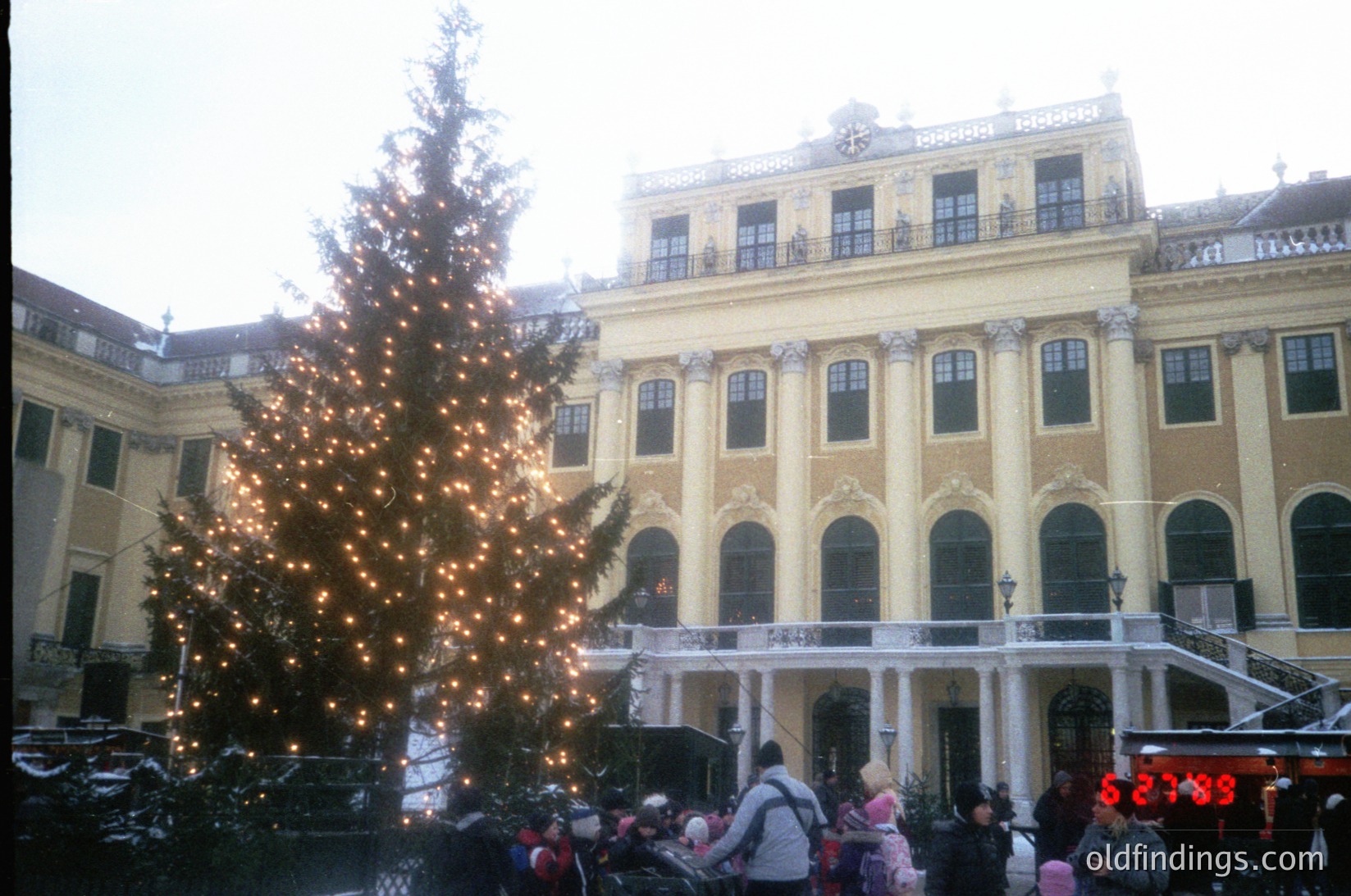 Neoclassical palace facade adorned with Baroque detailing, featuring arched windows and decorative cornices. A large, illuminated Christmas tree stands prominently in the courtyard, surrounded by festive crowds. Snow dusts rooftops and ground, indicating winter. Likely Vienna, Austria, 1980s–1990s.