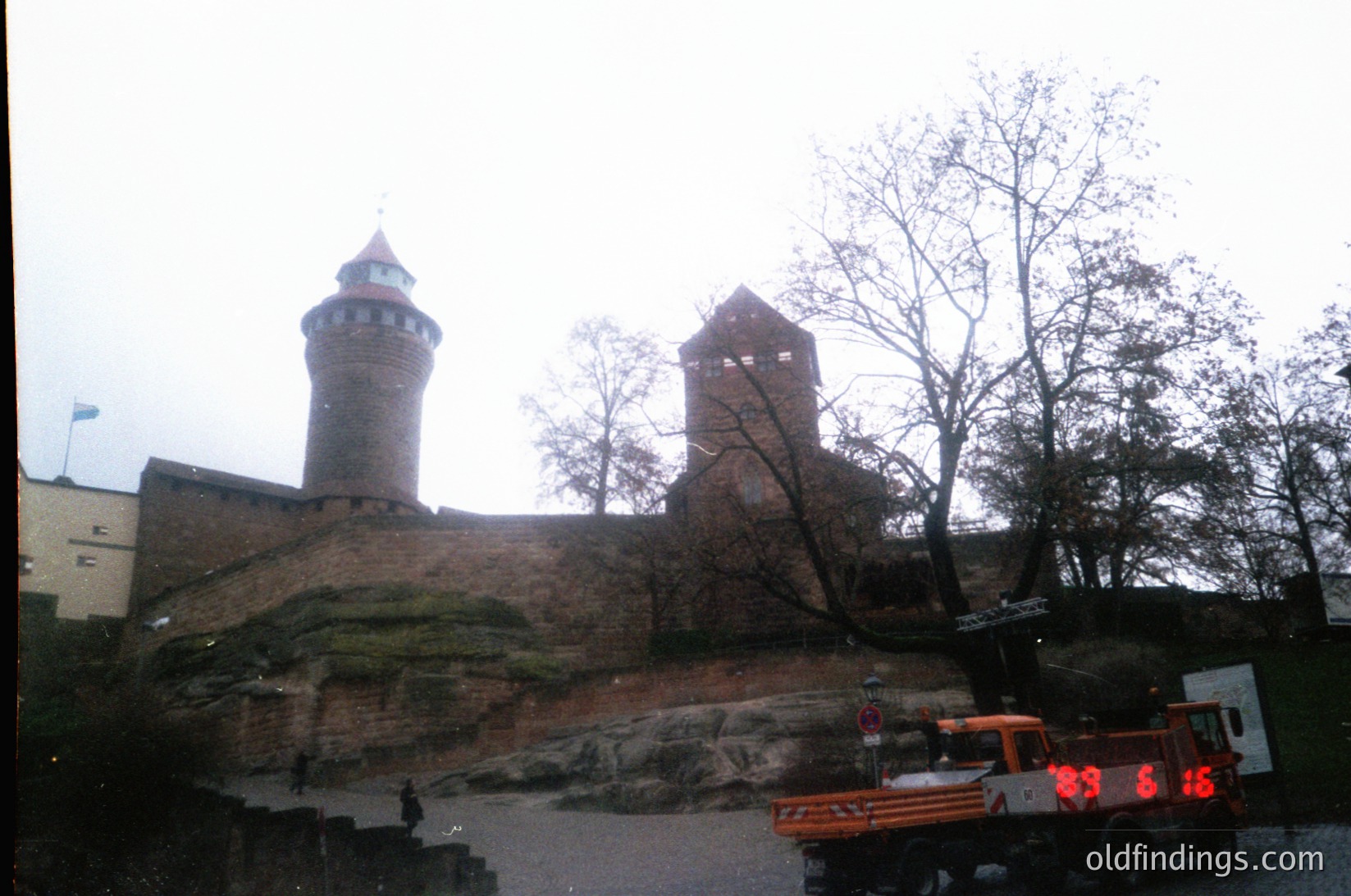 Medieval stone fortress with two cylindrical towers on elevated rocky terrain. Overgrown vegetation and a vintage truck (license plate "99 6-16") in foreground. Likely Eastern European, 1990s.