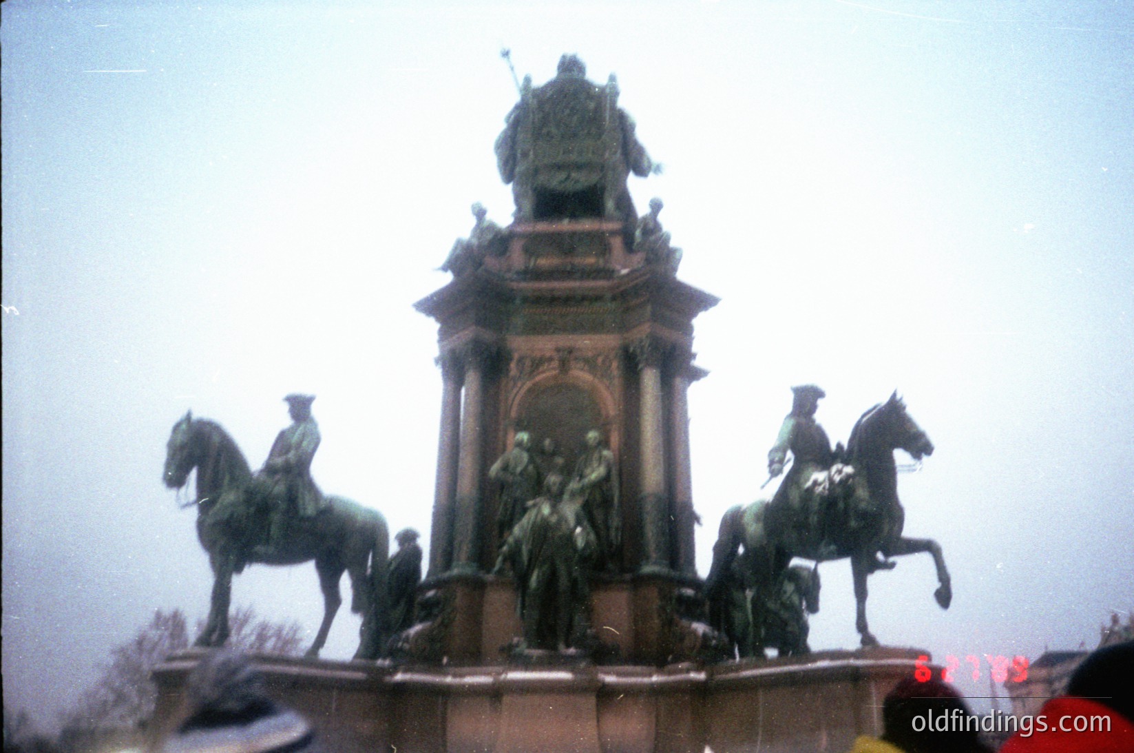 Neoclassical equestrian monument featuring three mounted figures flanking a central seated group atop an ornate pedestal. Intricate bronze detailing and architectural motifs suggest 19th-century European design. Fog obscures background, emphasizing monument’s prominence. Likely a public square or plaza.