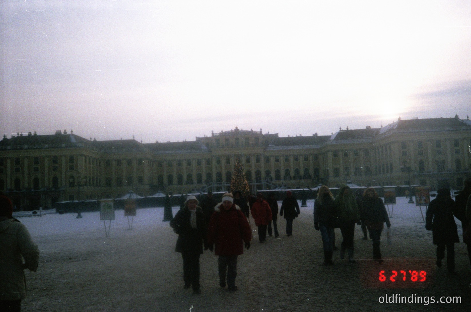 Neoclassical palace courtyard in winter, likely **Schönbrunn Palace, Vienna, Austria**. Frost-covered ground and low winter light frame grand colonnades. Crowd in 1980s winter attire (coats, scarves) strolls toward illuminated central fountain. Architectural symmetry and historic grandeur evident. önbrunnPalace