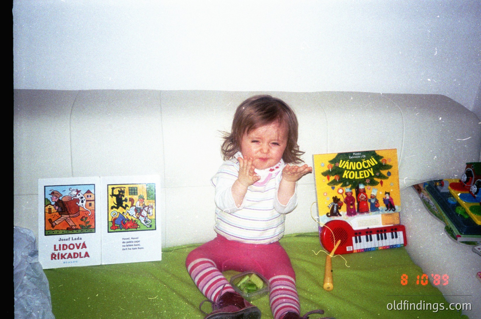 A toddler sits on a green play mat in a 1990s indoor play area, clapping hands. Surrounding them are Czech children’s books: *"Lidová říkadla"* (folk sayings) and *"Vánoční koledy"* (Christmas carols), alongside a toy piano. The date stamp reads **8.10.99**.
