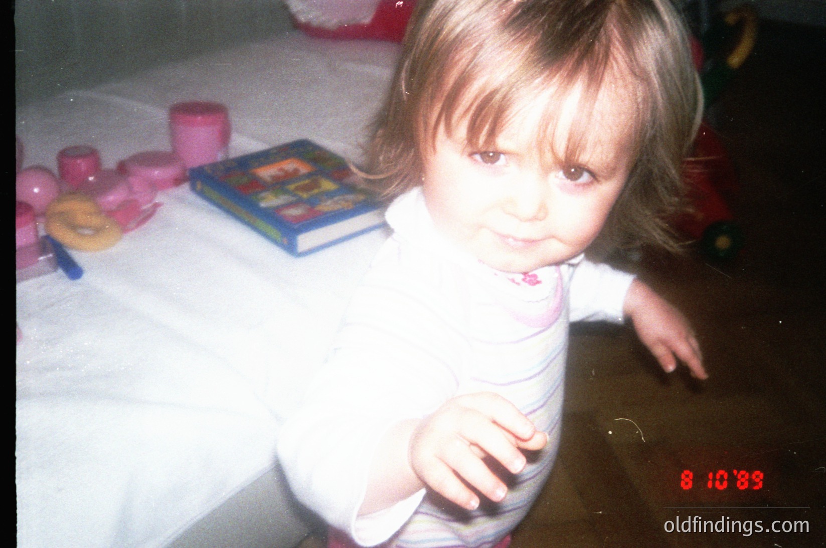 Vintage indoor portrait of a young child (approx. 2-3 years) seated at a table with 1990s-era toys: pink plastic cups, a toy phone, and a board game. Warm lighting and dated film grain suggest late 1990s. Candid, unposed expression captures innocence.