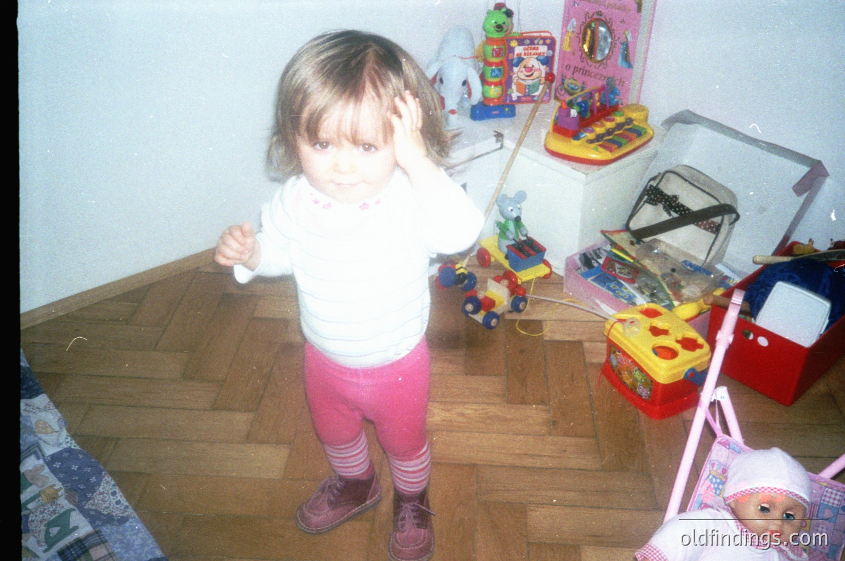 Young child in 1980s/90s indoor playroom, holding toy steering wheel. Surrounding toys include a toy piano, colorful plastic construction set, and a doll with a pink bonnet. Hardwood floors and vintage wallpaper suggest mid-century home decor.