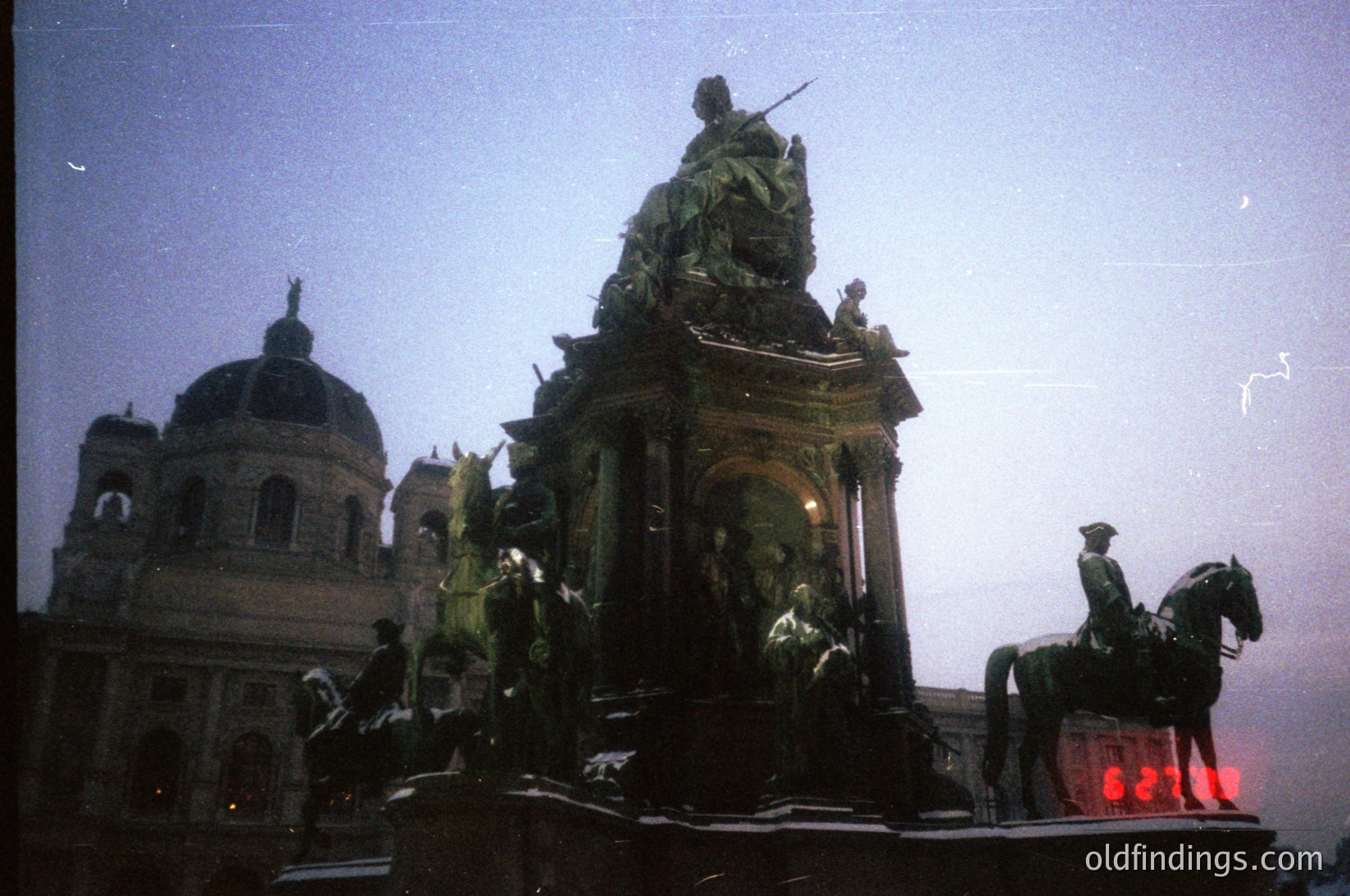 Equestrian statue and grand equestrian monument in dusk lighting, featuring a central figure atop a horse with surrounding figures. Behind it, a domed building with intricate architectural details. Likely Budapest, Heroes' Square, 19th-century Hungarian Revival style.