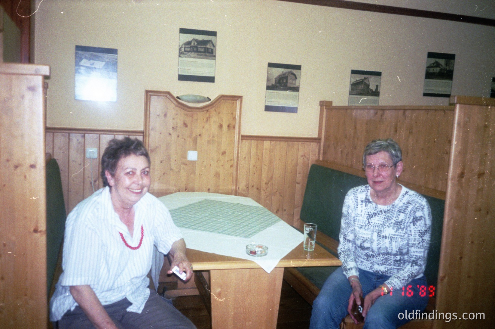 Two women seated at a wooden café table in a rustic wooden-paneled room, dated 1985. The woman on the left wears a white blouse with a red necklace, while the right wears a patterned sweater. A green map and empty glasses rest on the table. Framed scenic postcards adorn the walls, suggesting a rural or alpine setting. Warm, vintage lighting enhances the nostalgic atmosphere. éCulture