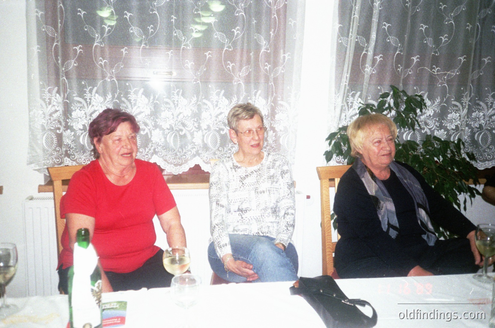 Three women seated indoors at a table, likely in a domestic setting. The woman on the left wears a red top, the center a patterned blouse with jeans, and the right a dark top with a scarf. A glass of wine and a bottle of water sit on the table. Floral curtains and a potted plant frame the scene. Style suggests mid-to-late 20th century. [Vintage family portrait ]