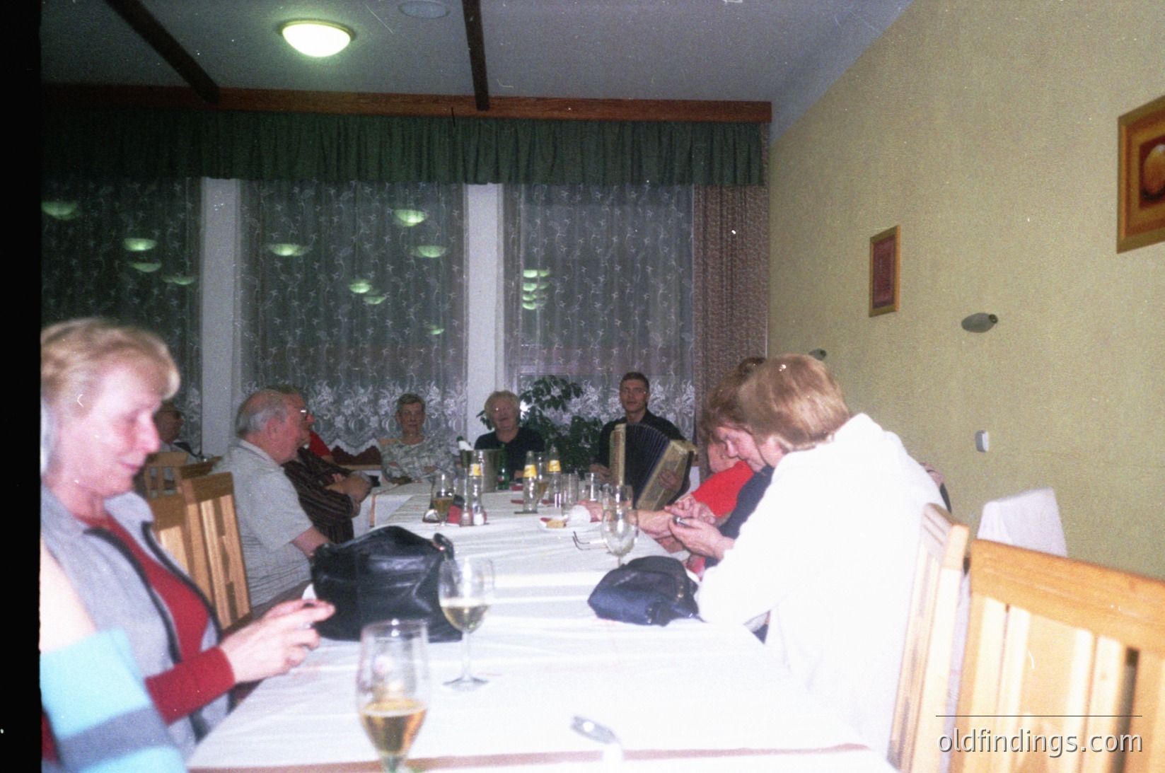 Vintage indoor gathering at a long dining table with 8 adults seated, dressed in 1980s-90s casual attire. Glassware, wine bottles, and a floral centerpiece suggest a social meal. Dim lighting and patterned curtains hint at a cozy, possibly residential dining area.
