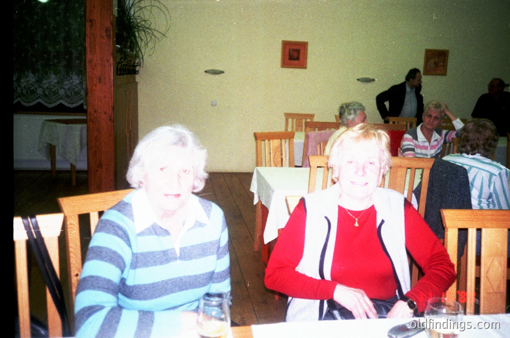 Vintage indoor café scene with two women seated at a wooden table, likely or . The woman on left wears a striped sweater, while the woman on right dons a red sweater with black stripes. Wooden chairs, framed art, and a potted plant decorate the warm-toned room.