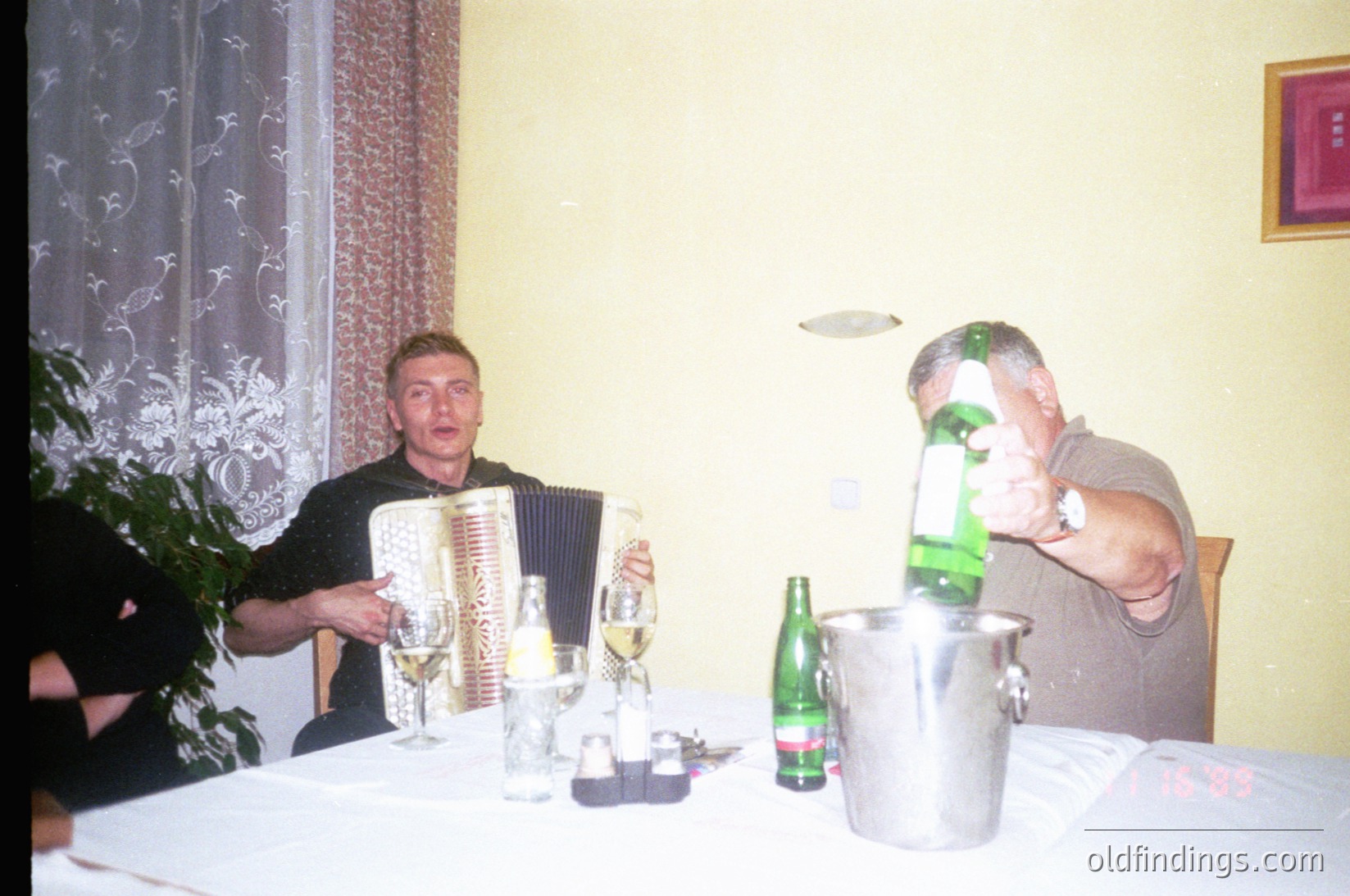 Intimate gathering featuring two men at a table: one playing an accordion, the other pouring beer from a bucket. Table set with wine glasses, beer bottles, and a potted plant. Decor includes lace curtains and a framed painting. Likely a private celebration or folk event, Eastern European setting, 2000s.