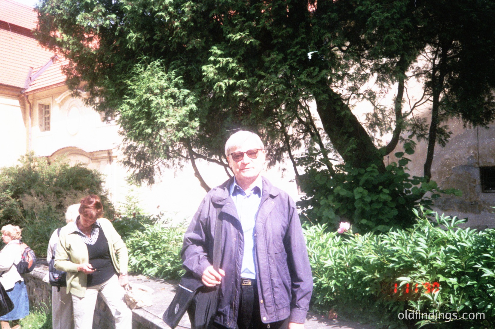 Two adults in 1970s-style attire pose outdoors in a lush, shaded garden. The man wears a dark jacket, sunglasses, and a light shirt; the woman in the background wears a light jacket and dark pants. Stone walls and greenery frame the scene, suggesting a historic or institutional setting. The timestamp "1979" is visible in the corner.