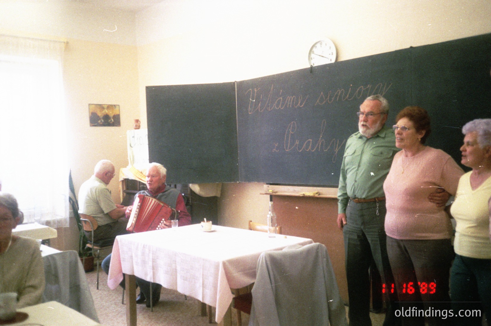 Vintage indoor gathering in a classroom setting, dated **1983**. Six adults—four men and two women—engage in a folk music session. A man plays an accordion at a table draped with a white cloth, while others stand or sit, some clapping. Blackboard text reads *"Vítáme seniory z Brahy"* (welcoming seniors from Braha). Simple wooden chairs, a clock, and framed artwork decorate the room.