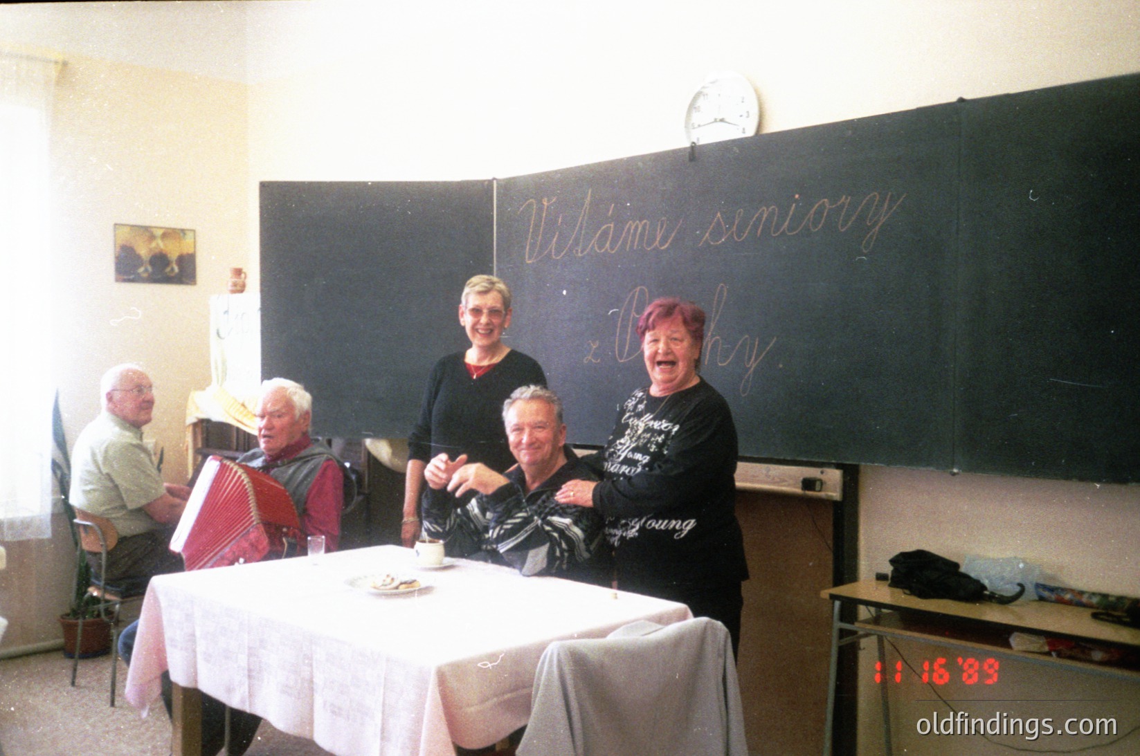 Vintage classroom scene with chalkboard reading *"Vitáme seniory"* ("Welcome seniors") in Czech. Five adults pose around a table with a white tablecloth, likely from a 1980s–1990s Czech or Slovak senior gathering. One woman wears a "Young" t-shirt, suggesting a generational mix. Clock shows 11:15.