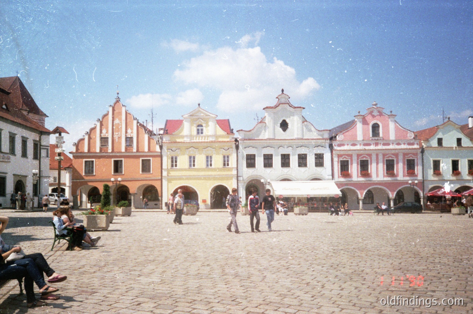 Colorful Baroque-style townhouses line a cobblestone square, featuring pastel facades (). People casually stroll or sit on benches under clear skies. Likely a European town square, possibly or , mid-20th century ().