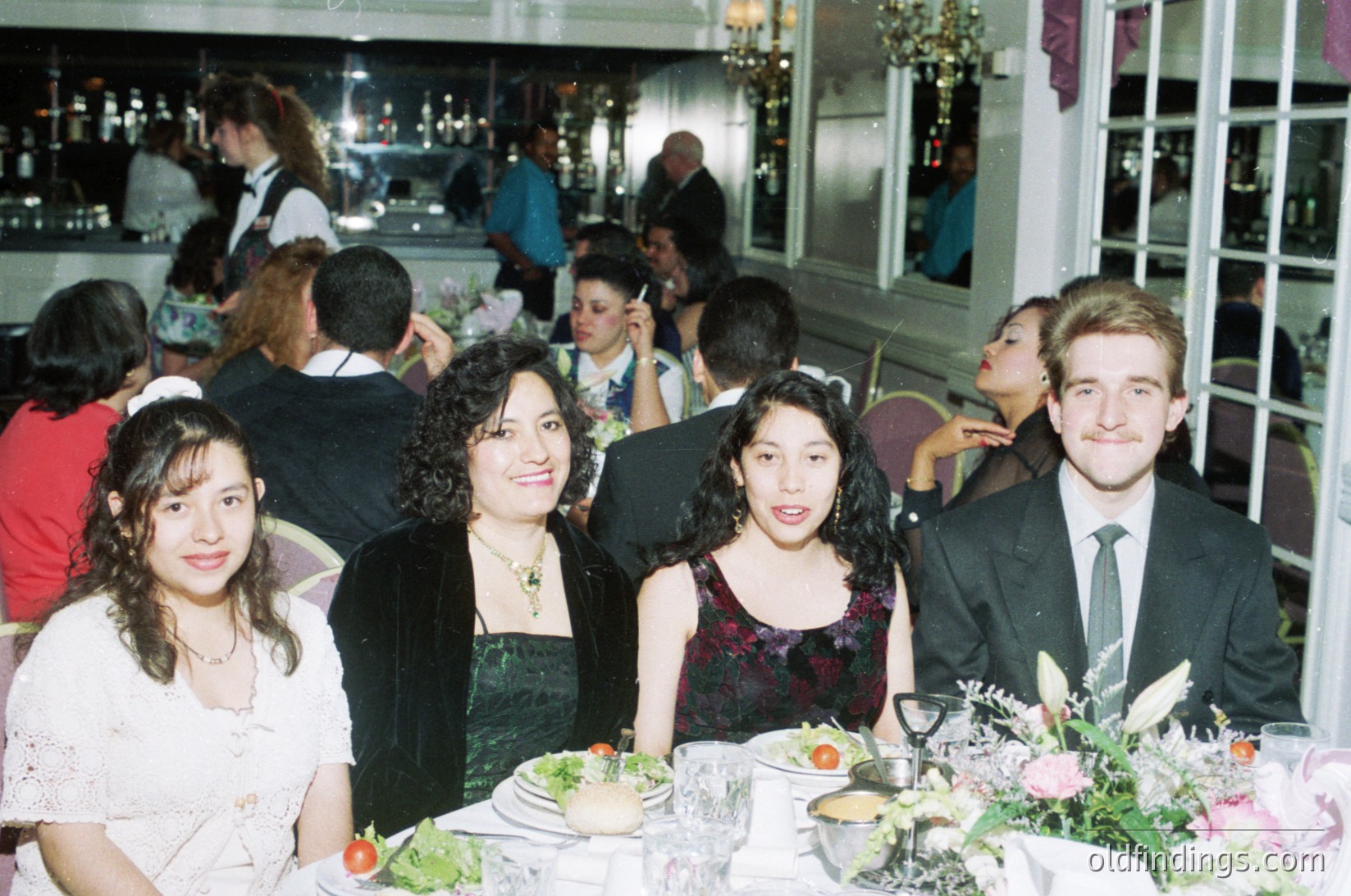 Vintage group portrait at an elegant evening event, likely a wedding or formal reception. Four individuals pose at a table adorned with floral centerpieces and wine glasses. The woman on the left wears a white lace dress, while others in formal attire—dark dresses and suits—complement the sophisticated setting. Blurred background reveals a crowded hall with chandeliers and guests, suggesting a mid-to-late 20th-century venue.