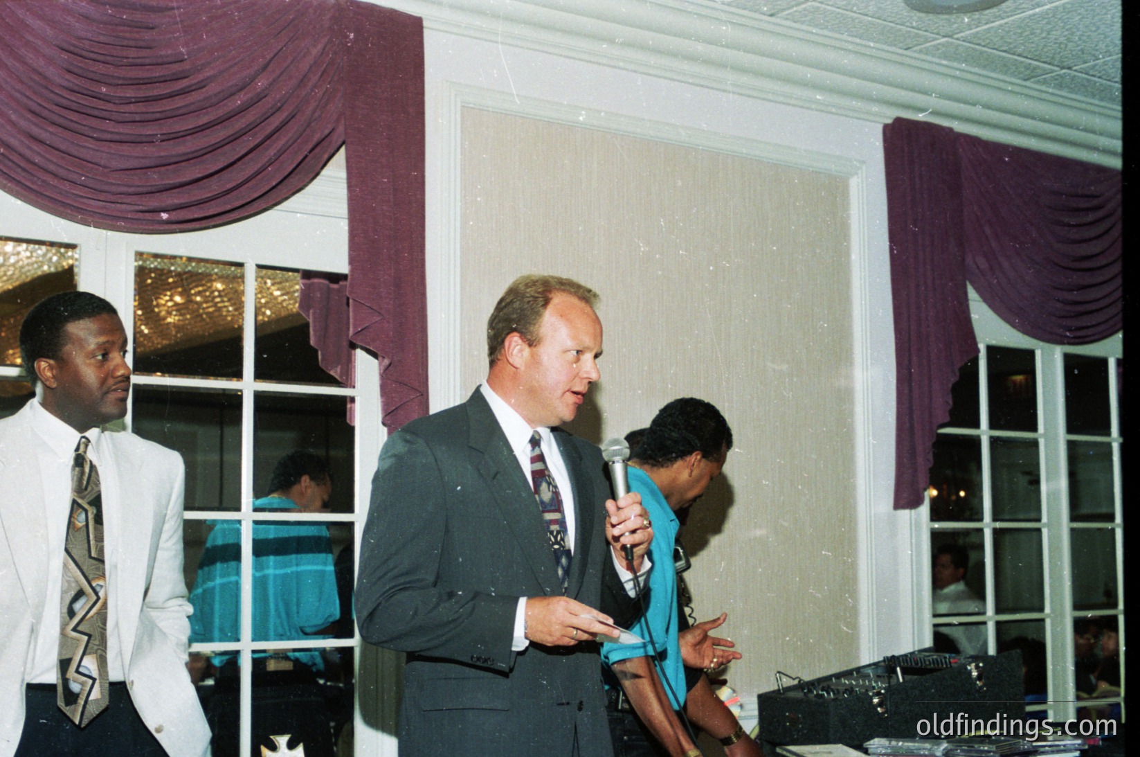 Formal indoor event with three men in 1990s business attire—dark suits, ties, and dress shirts. Central figure holds a microphone, speaking at a podium. Purple drapes frame large windows, revealing a blurred outdoor scene. Decorative tablecloth and scattered event materials suggest a conference or seminar setting.