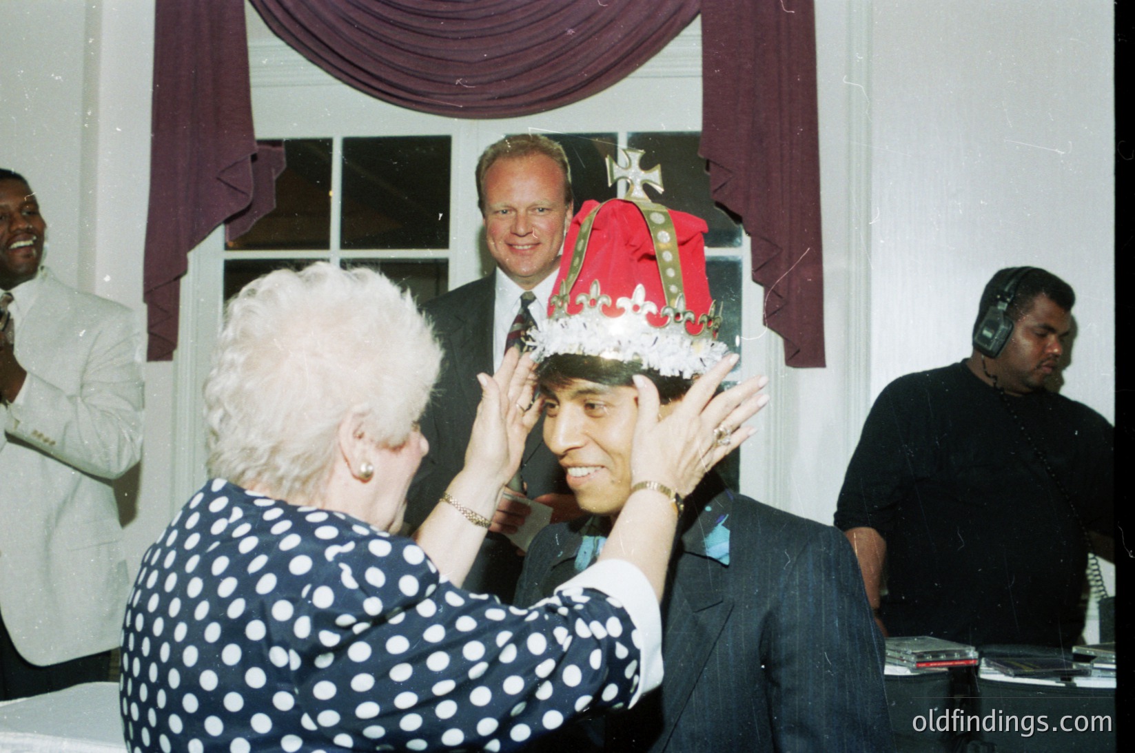 A formal event featuring a ceremonial crowning. Elderly woman in a polka-dot dress places a red, gold-embellished crown on a man’s head, supervised by a man in a suit. Background includes draped red fabric and a window. Likely a cultural or community celebration, possibly