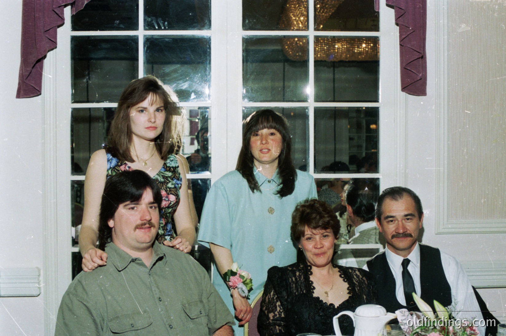 Five adults pose indoors near a large window with decorative trim, likely a 1980s-1990s wedding or formal event. Men wear dress shirts with ties/vests; women in floral/patterned blouses with lace details. Center table displays floral arrangements and teacups.