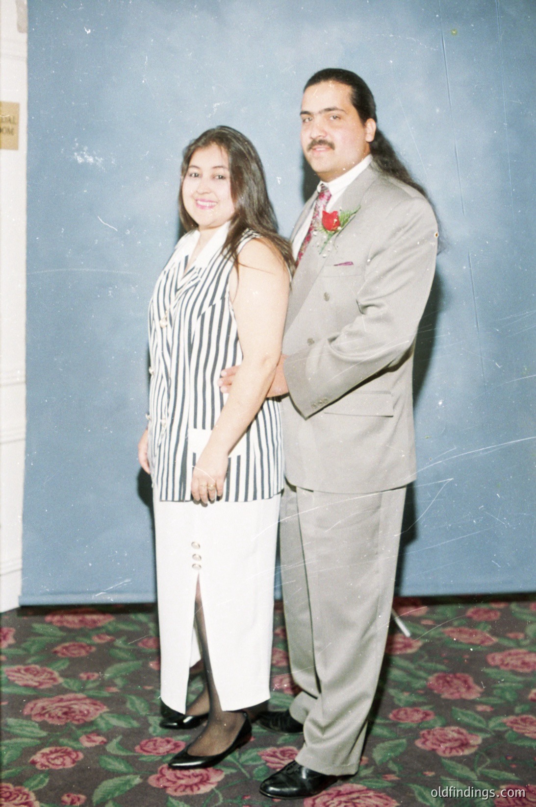 A 1990s-era formal portrait featuring a man and woman in coordinated attire. The man wears a light gray suit with a red boutonnière, while the woman’s striped dress (black/white) complements his ensemble. Floral carpet and muted blue wall suggest an indoor venue, likely a wedding or formal event.