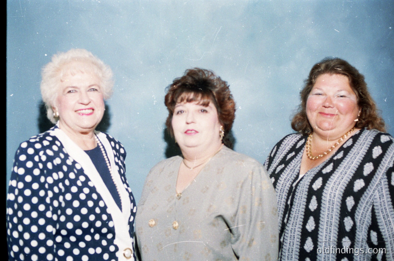 Three women pose formally in 1980s/90s attire—polka-dot blazer, cream blouse with buttons, and striped sweater with pearl necklace. Soft focus and muted lighting suggest an indoor event, likely professional or celebratory.